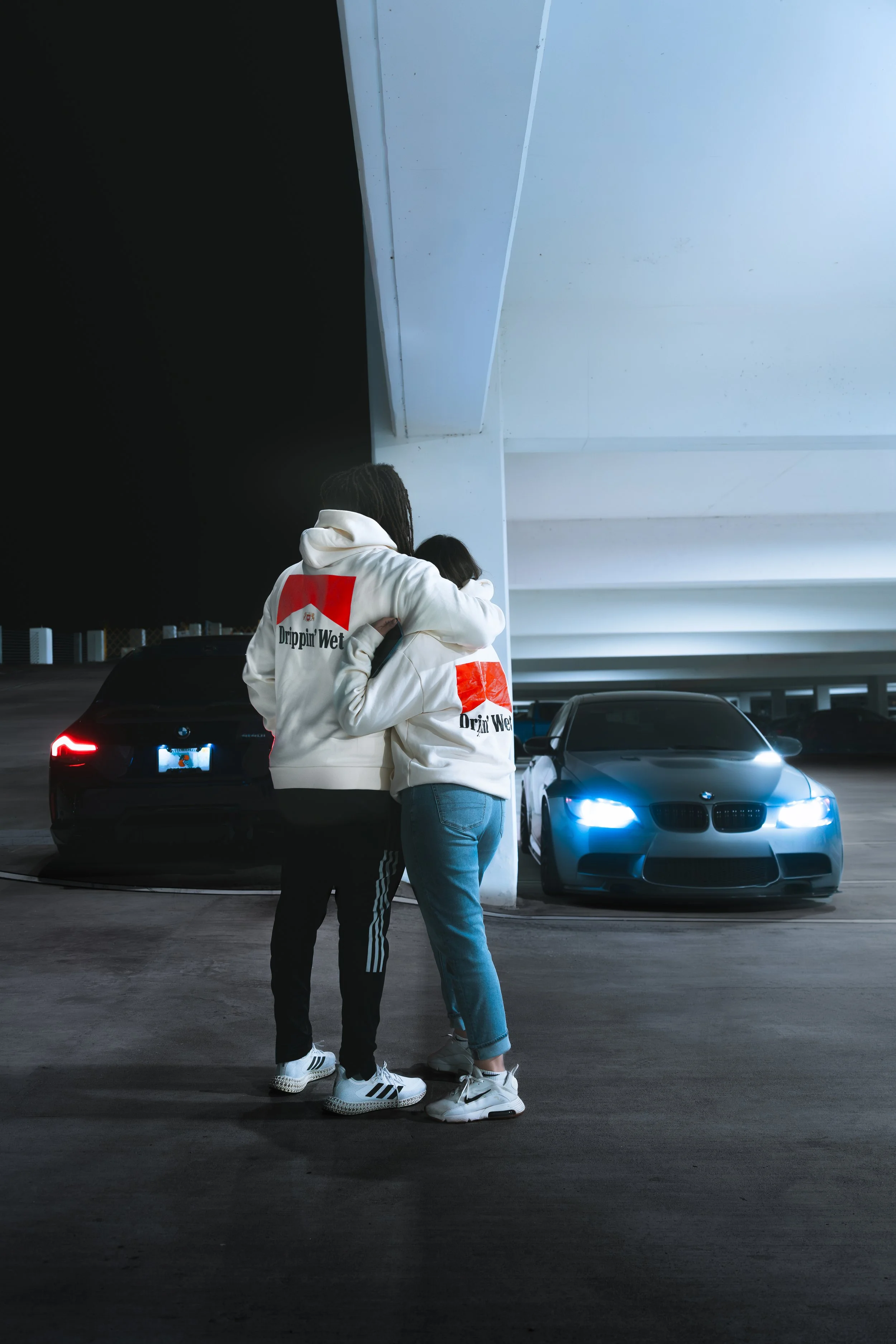 Two people hug in a parking garage at night, wearing white hoodies with red and black graphic designs. Their backs are visible, and there are modern cars with headlights on in the background.