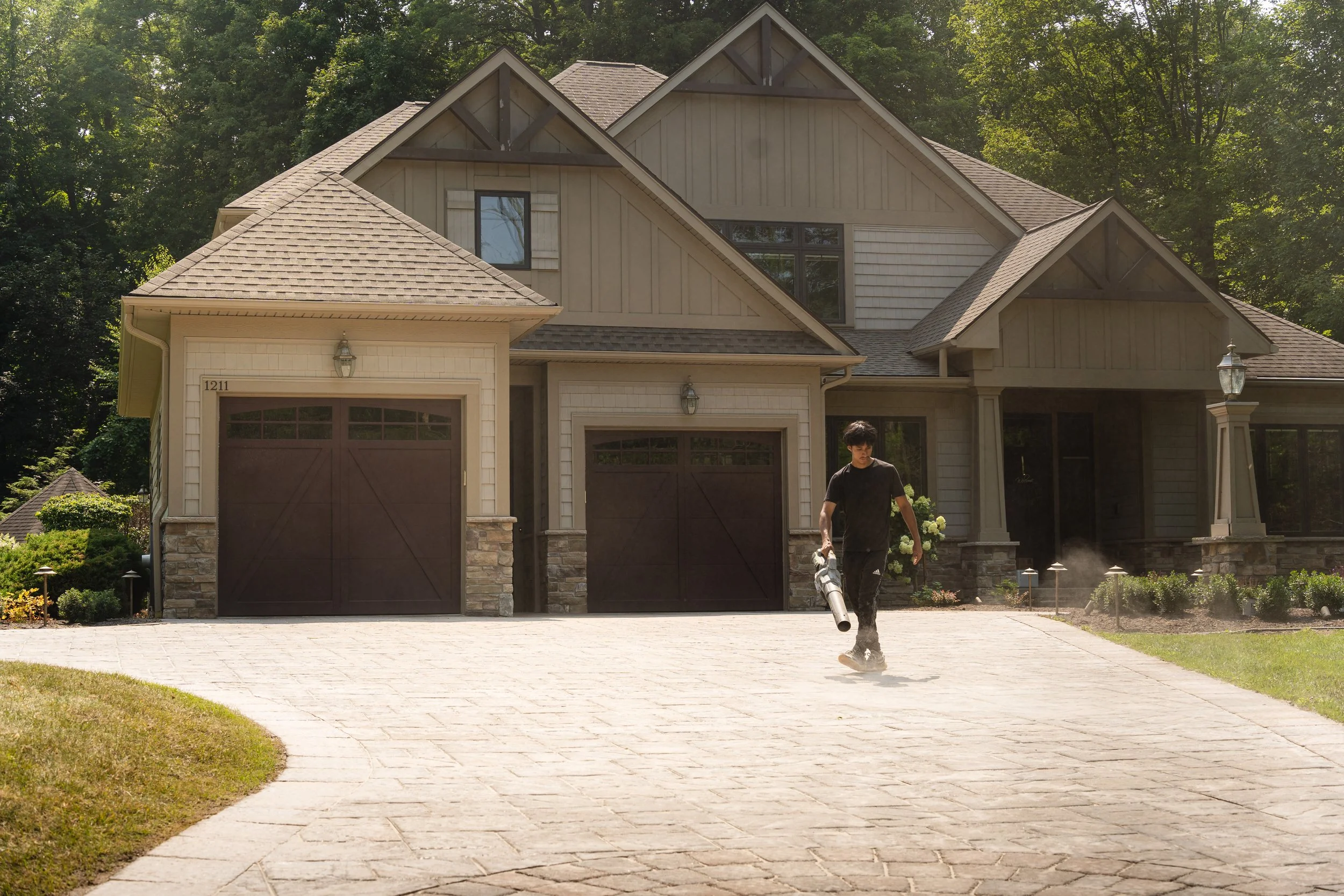 A person is using a leaf blower to clear dust or debris from a paved driveway in front of a large beige house with multiple gabled roofs and dark brown garage doors. The house is surrounded by greenery and trees.