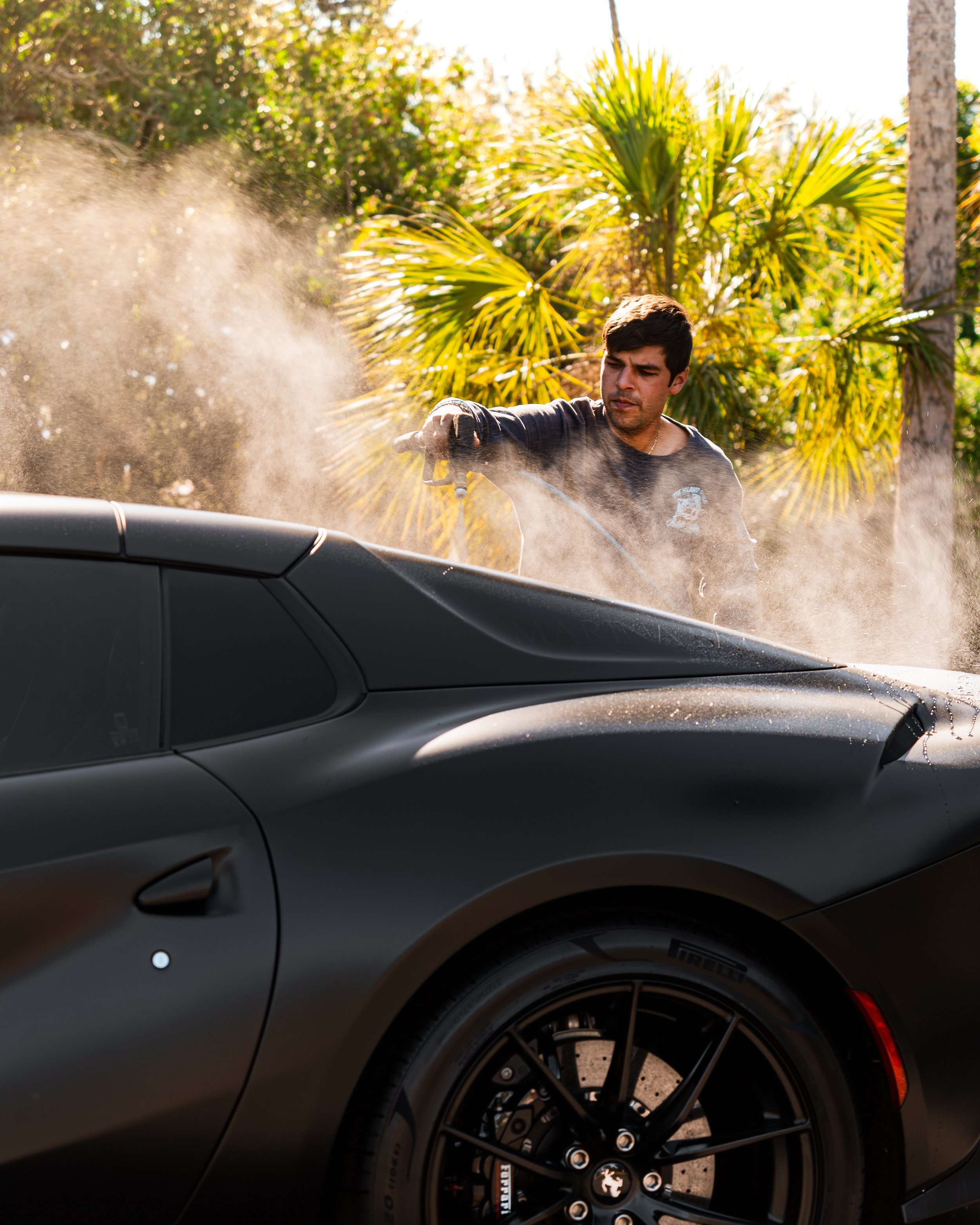 Man cleaning a matte black sports car outdoors amidst tropical plants.