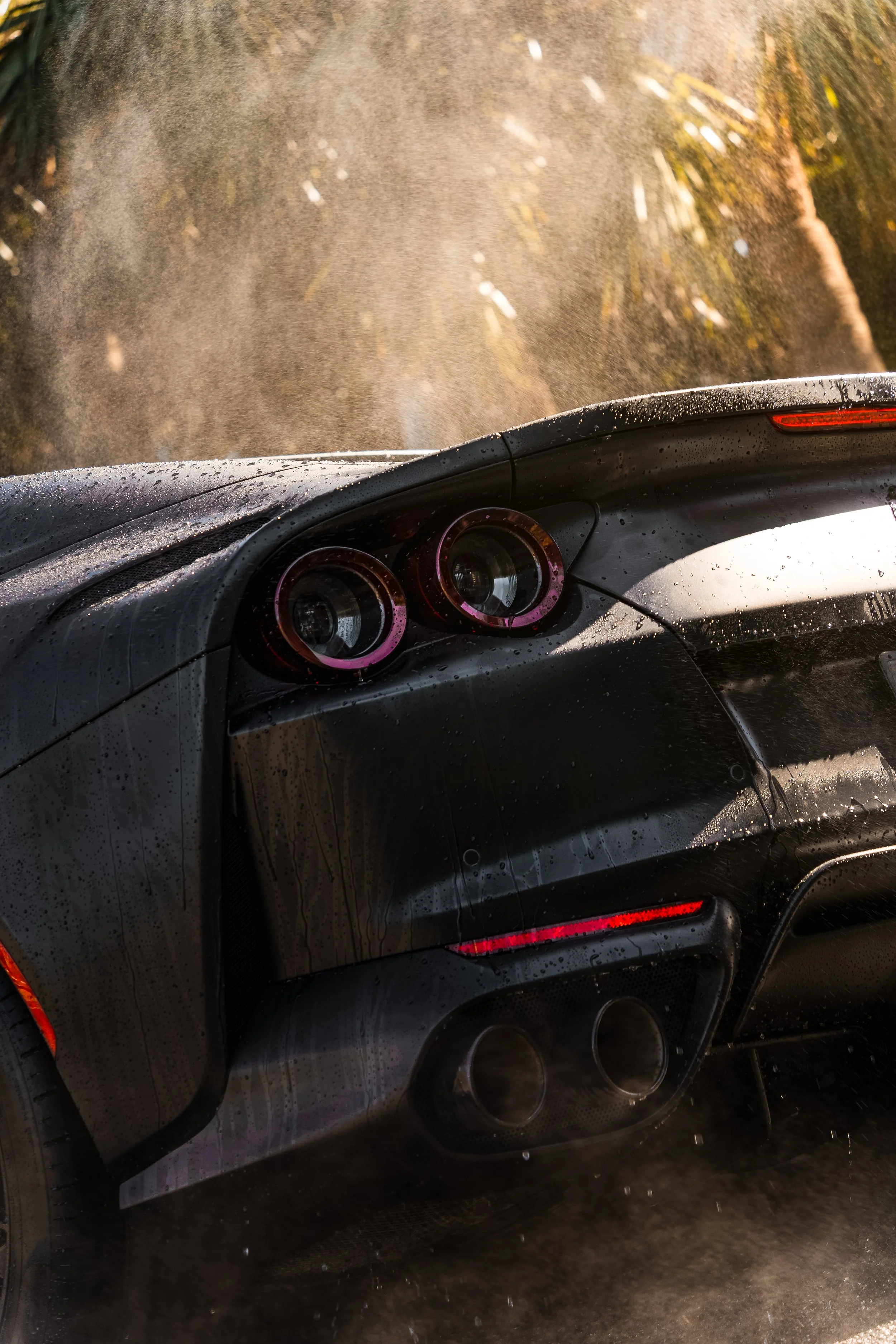 A black sports car with a sleek rear design, dual exhaust pipes, and water droplets on the surface, driving on a rainy road with trees in the background.