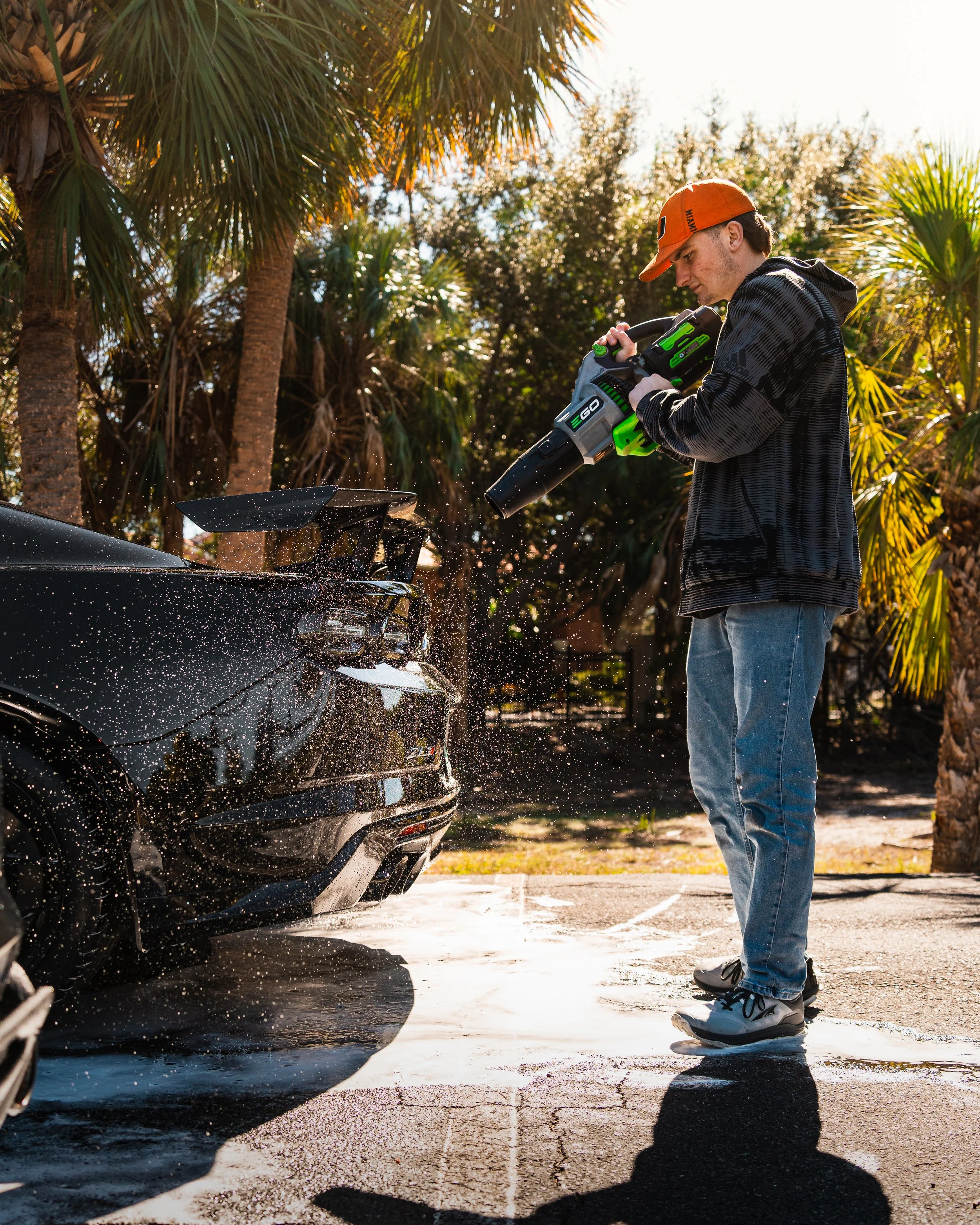 Man in a black hoodie and jeans using a leaf blower to clean a black sports car in an outdoor setting with palm trees in the background.