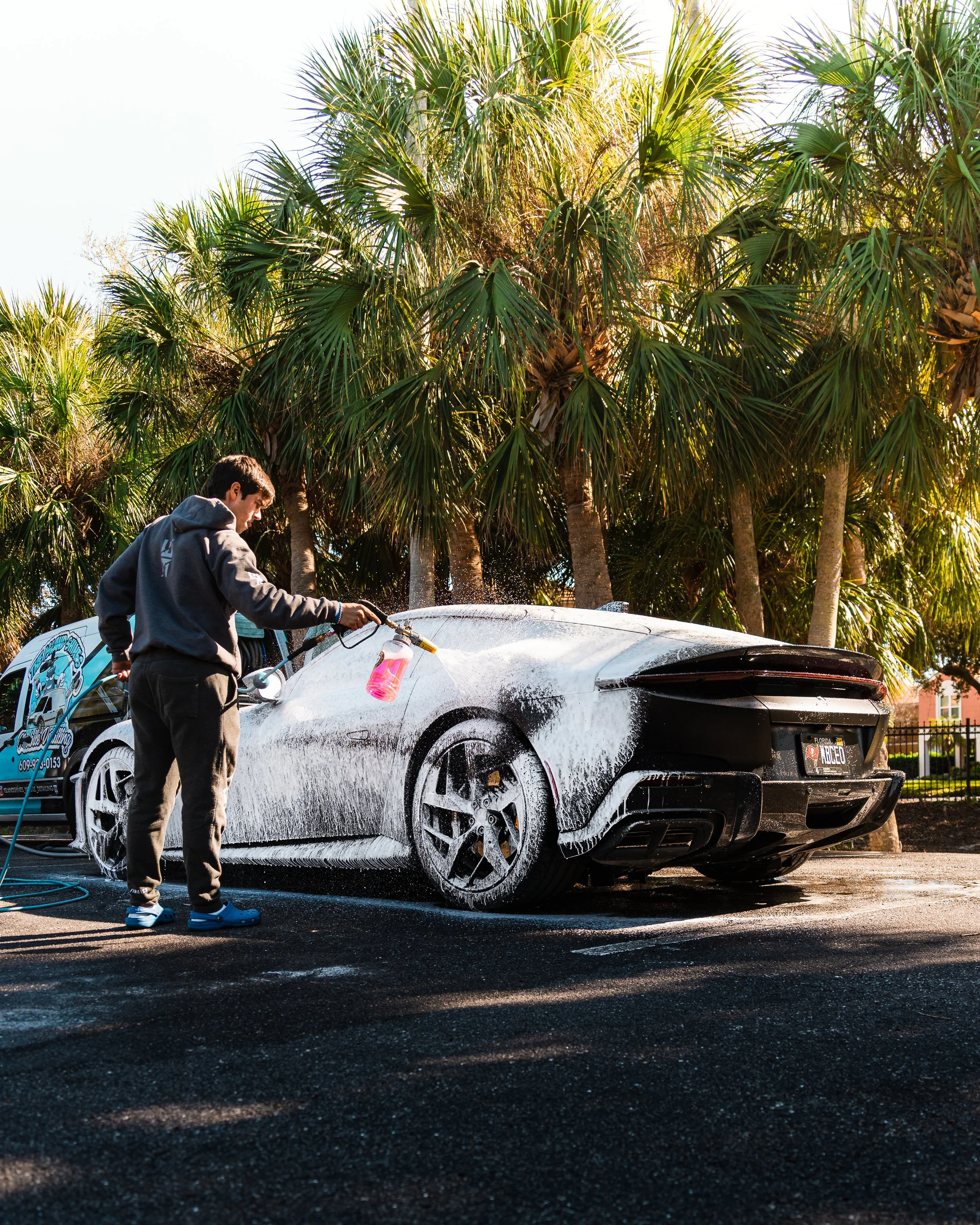 A man is washing a black sports car with soap and a foam cannon in an outdoor parking lot surrounded by tall palm trees.