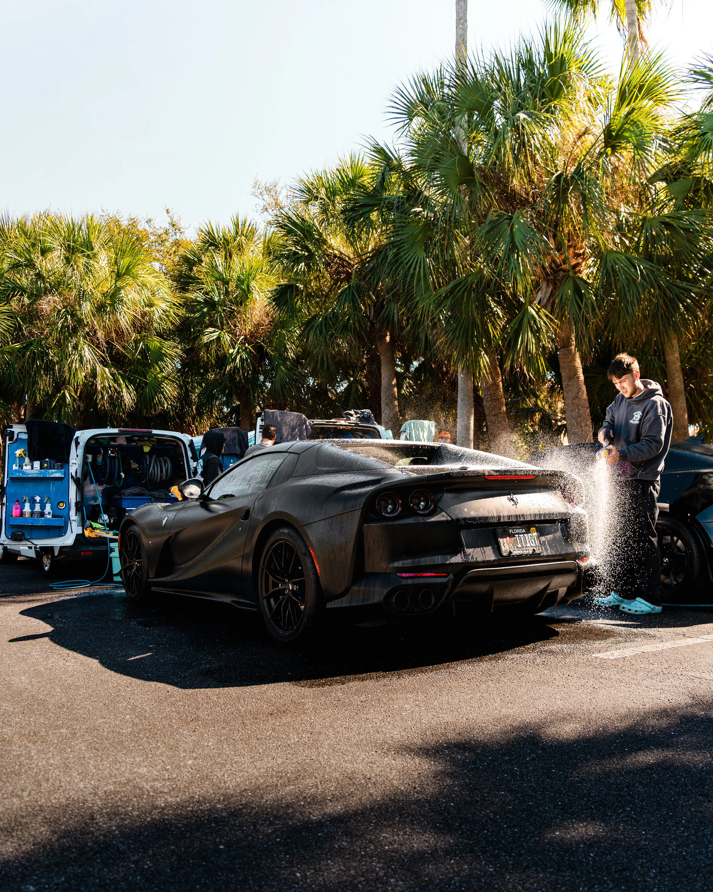 A person washing a matte black sports car in a parking lot surrounded by palm trees, with service trucks and other cars nearby.