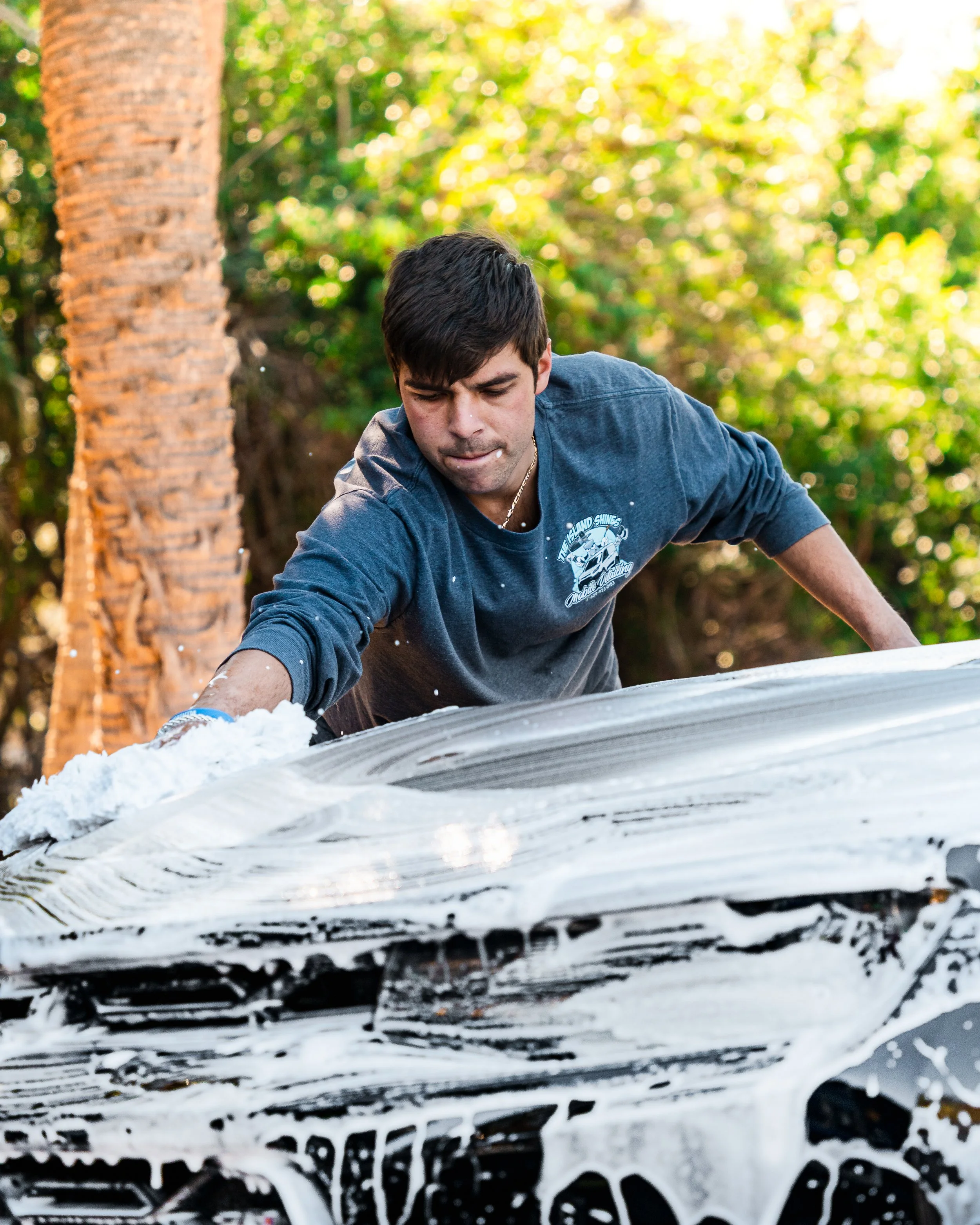 A man is washing a car with soap and water outdoors in a wooded area.