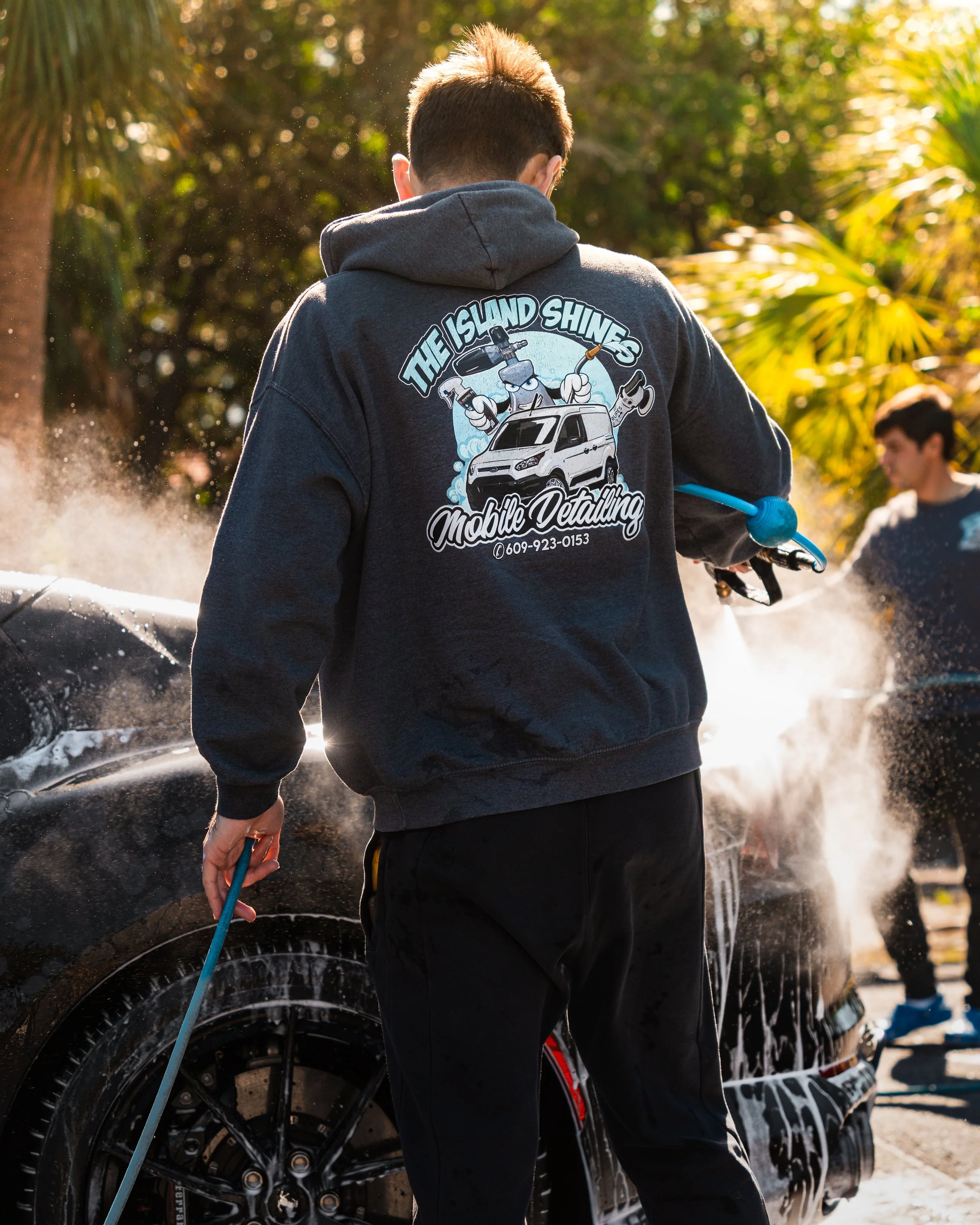 Car wash worker with a sweatshirt reading 'The Island Shines Mobile Detailing,' holding a hose, washing a black car with soap suds and water spray, with palm trees and another worker in the background.