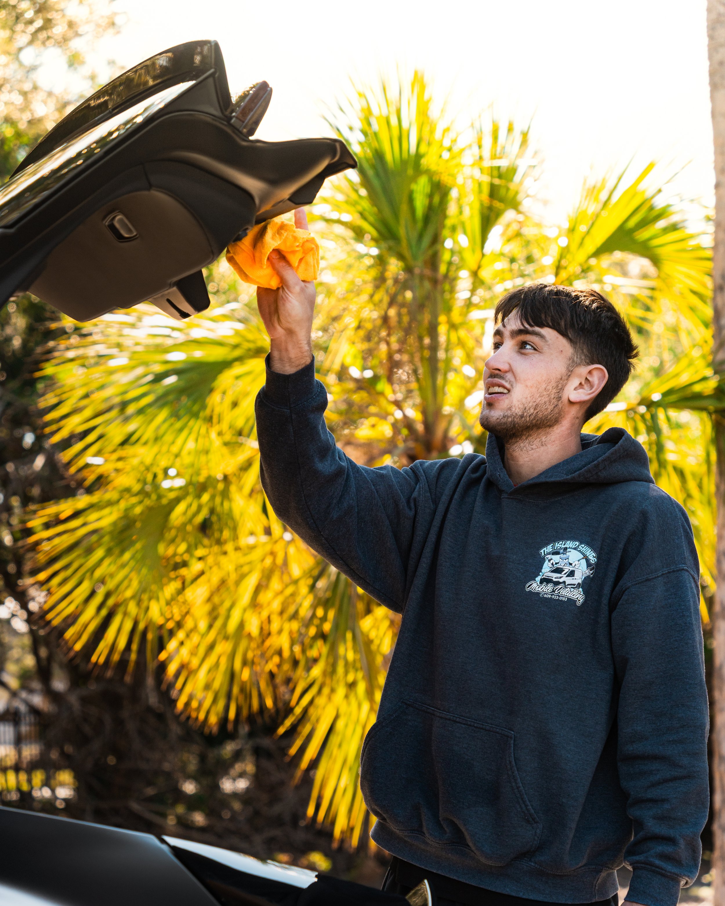 A young man cleaning a car windshield with a yellow cloth outdoors during daytime, with palm trees in the background.