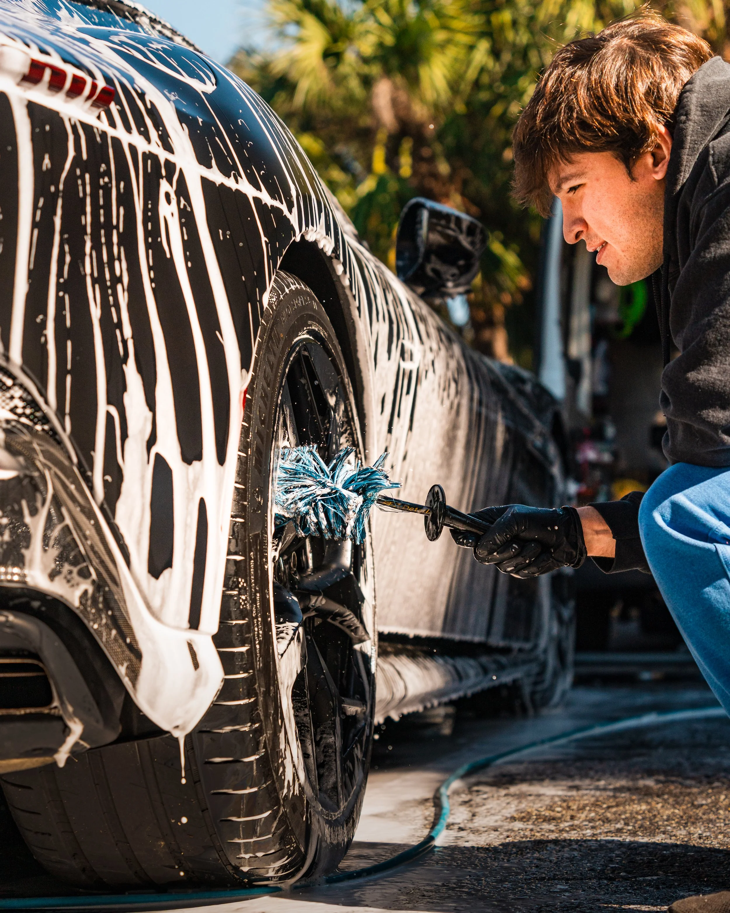 A person washing a black sports car with a foam cannon, creating soap suds and bubbles on the tire and body of the car during daytime.