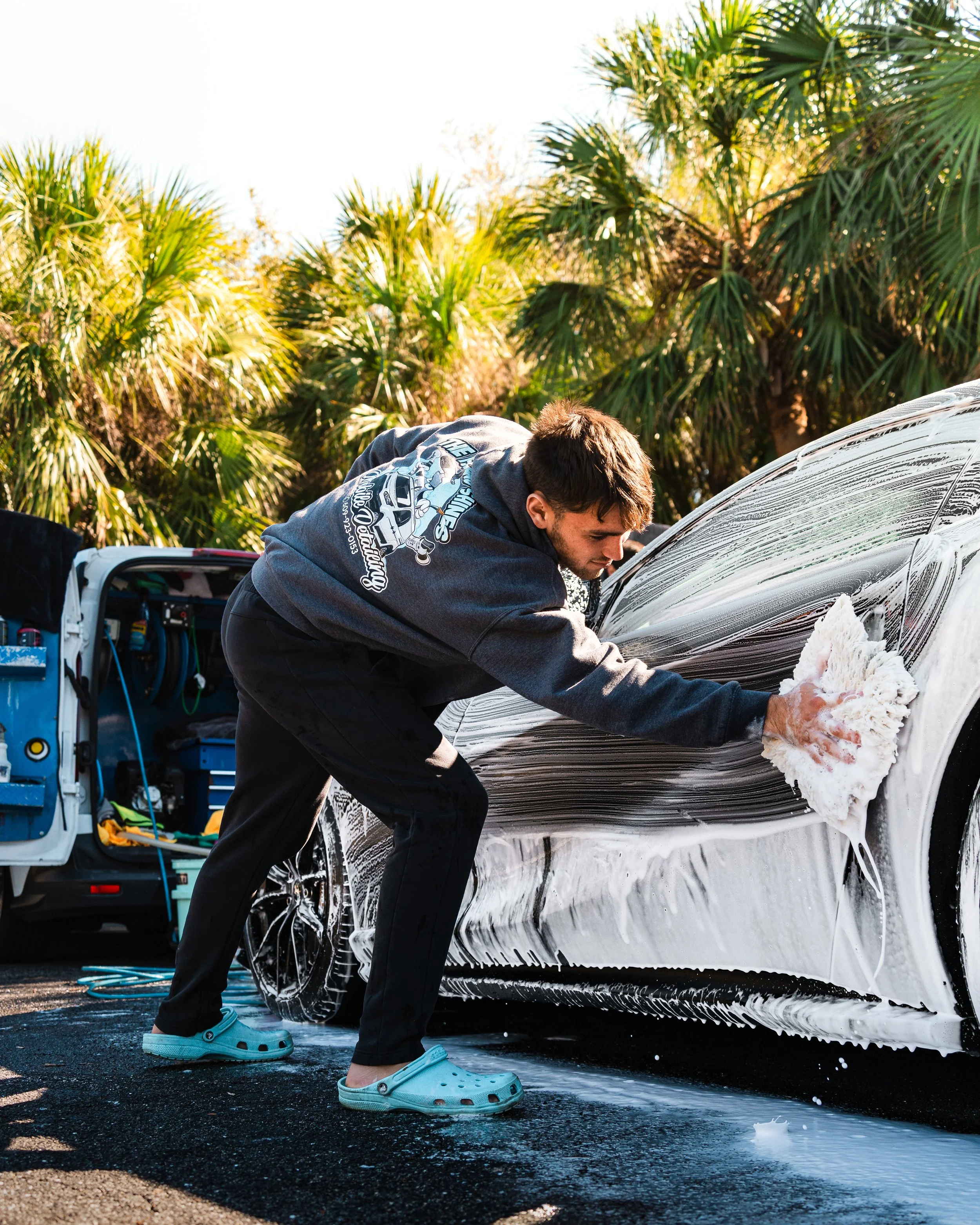 Man washing a black sports car with soap and sponge outdoors, palm trees in background, wearing blue Crocs and a dark hoodie.