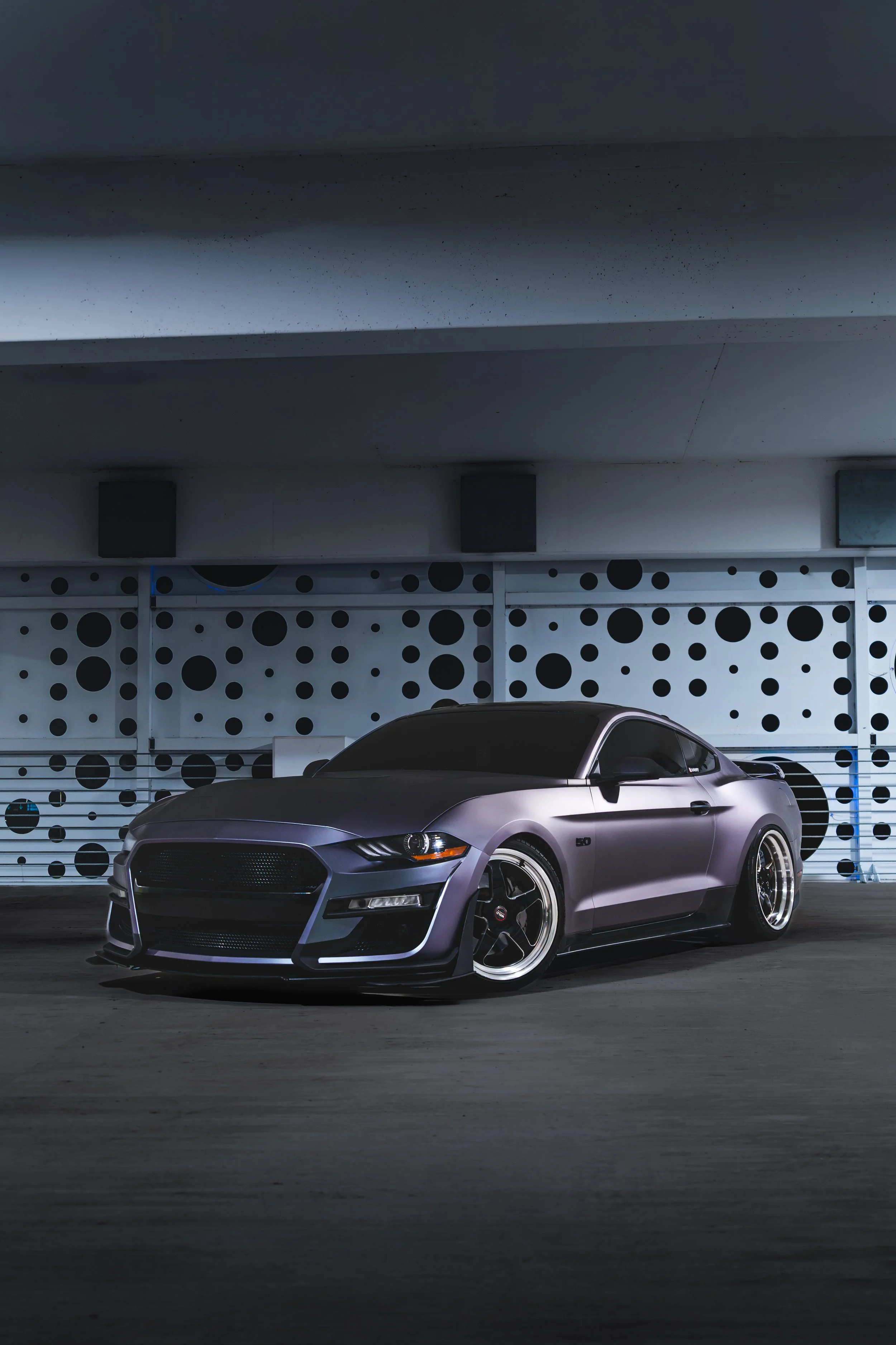 A sleek, matte gray sports car parked in an indoor parking garage with a black and white dotted wall in the background.