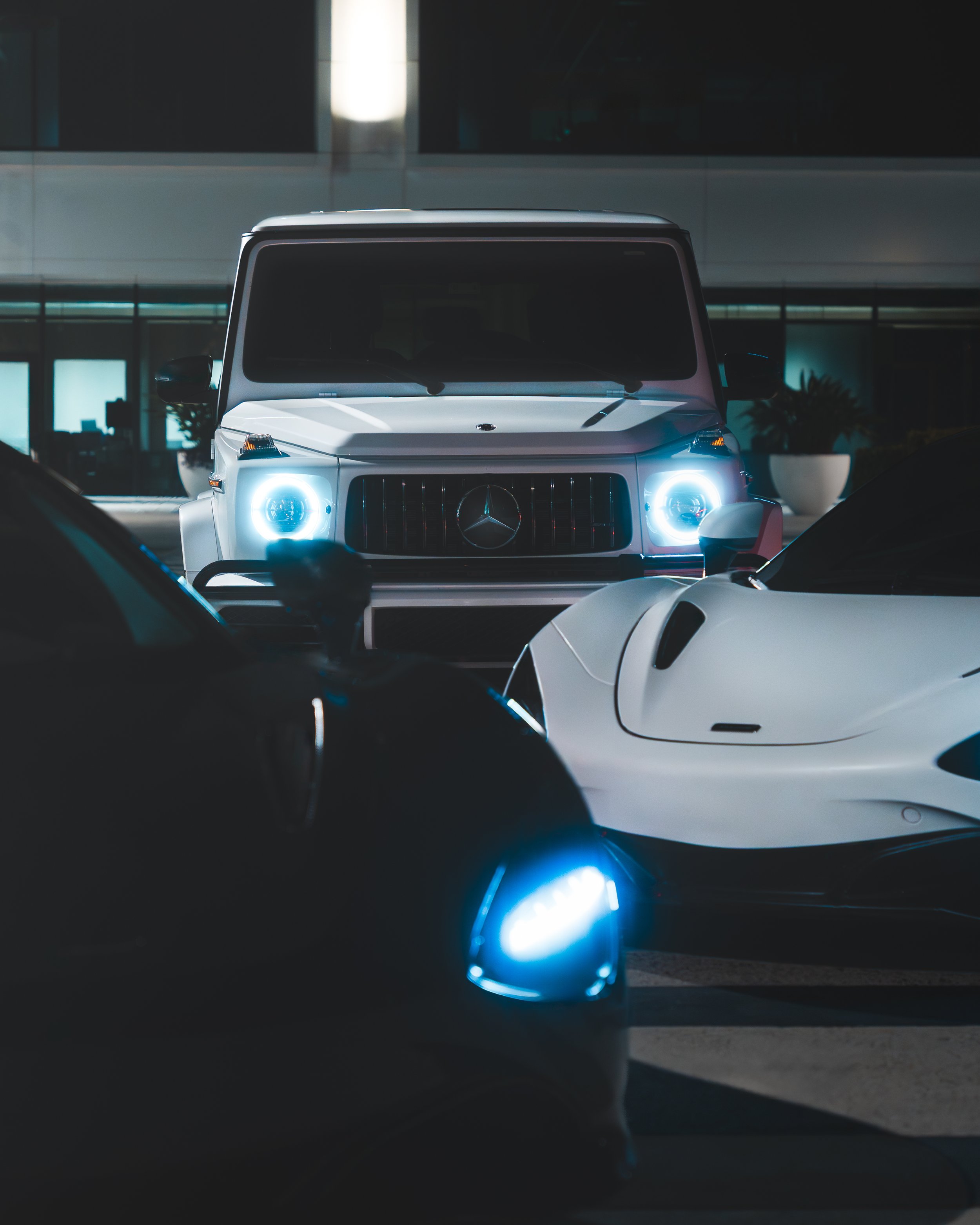 A white Mercedes-Benz G-Class SUV with illuminated headlights, flanked by two sports cars in front, one black and one white, parked indoors.