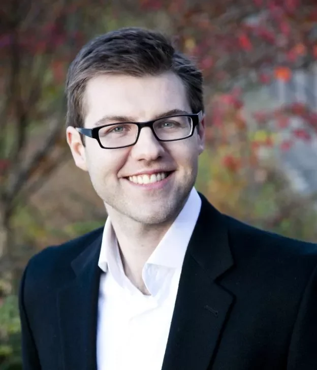 A smiling man with glasses, wearing a black blazer and white shirt, outdoors with blurred fall foliage in the background.
