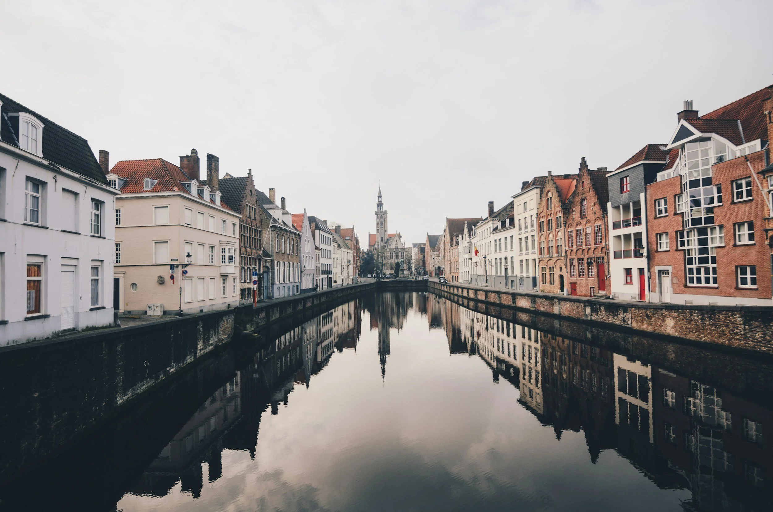 Cityscape of colorful European buildings lining a canal with a clock tower in the background under a cloudy sky, reflecting in the water.