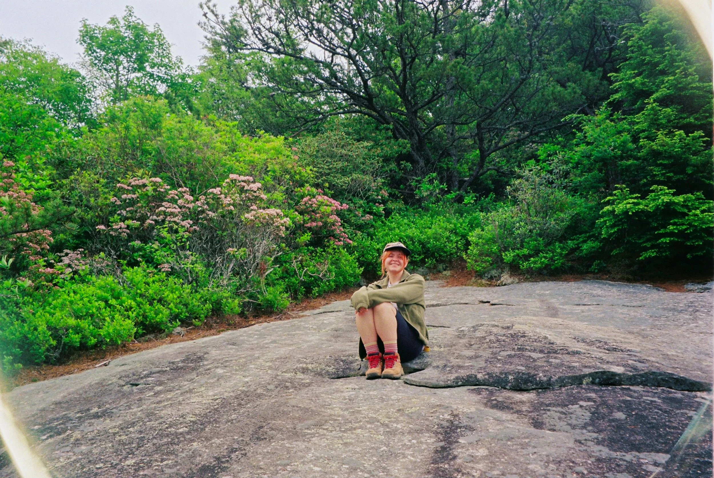 A young woman sits cross-legged on a large rock, smiling, surrounded by lush green bushes and trees in a natural outdoor setting.