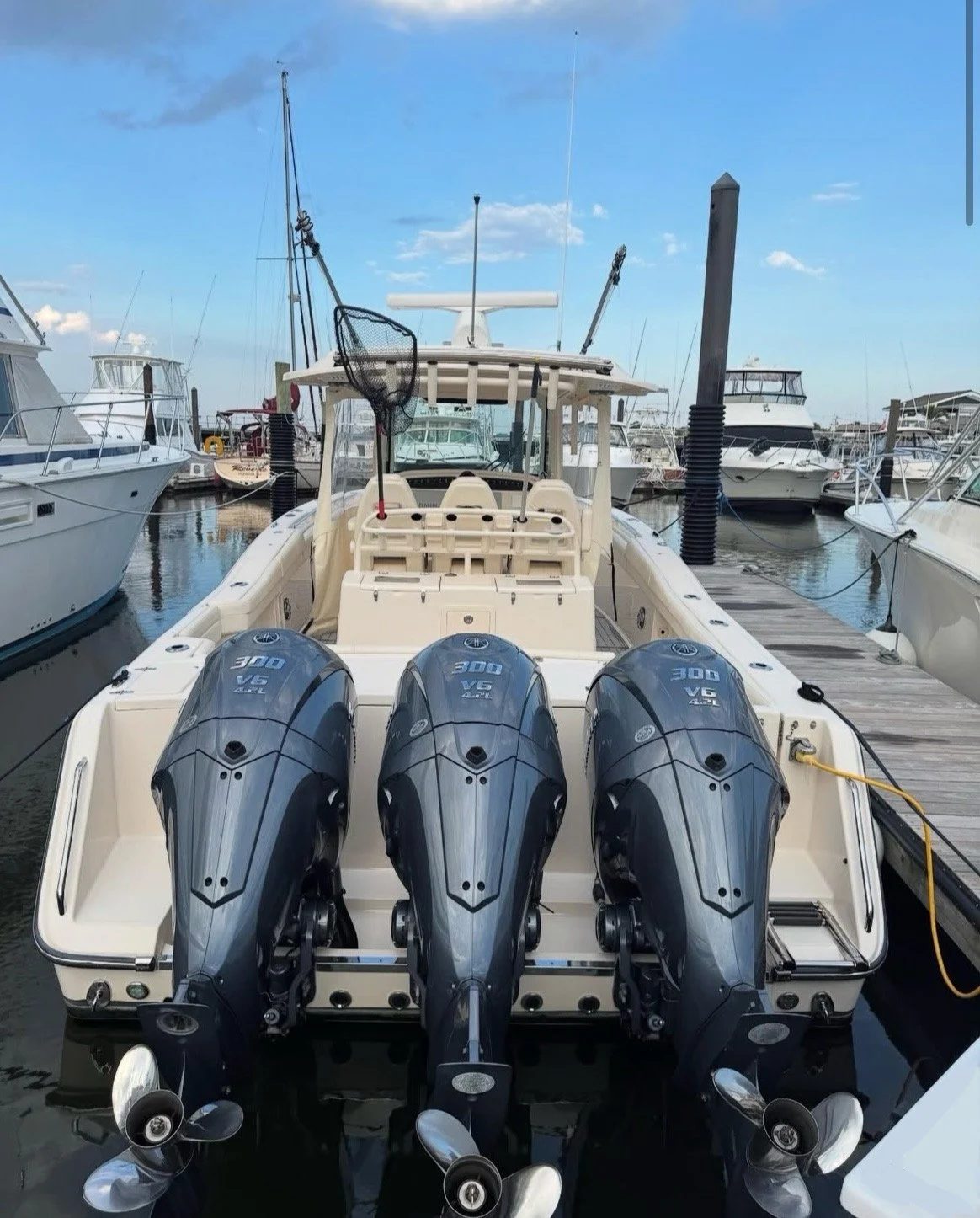 A boat with three outboard motors attached at the stern, docked at a marina with other boats and vessels on a clear day with some clouds