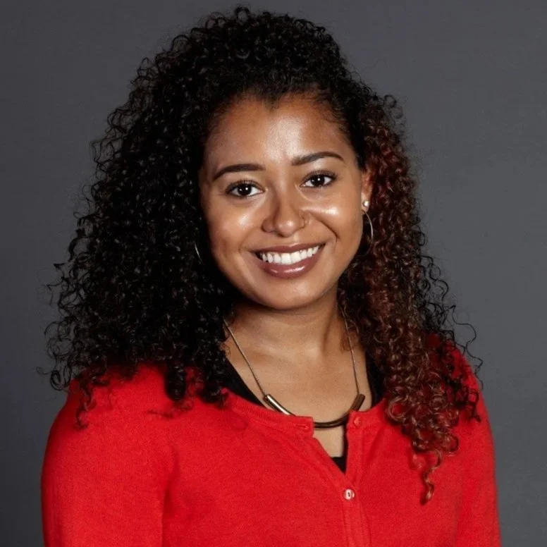 A woman with curly dark hair and brown skin smiling, dressed in a red top, standing against a dark gray background.