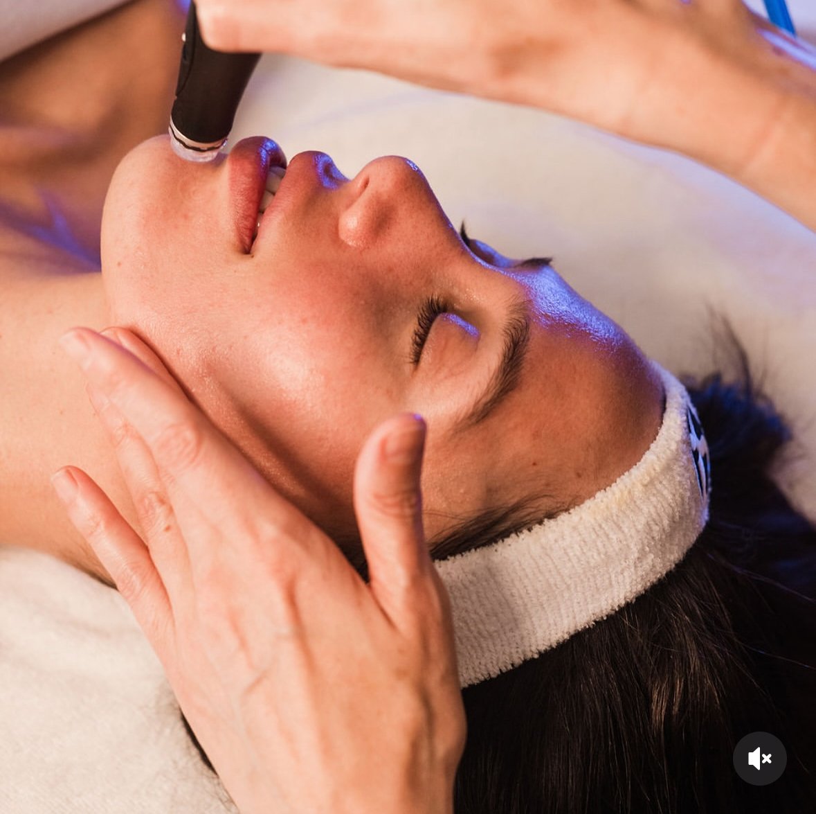 A woman receiving a facial massage at a spa, lying down with eyes closed, with a headband keeping hair away from face.