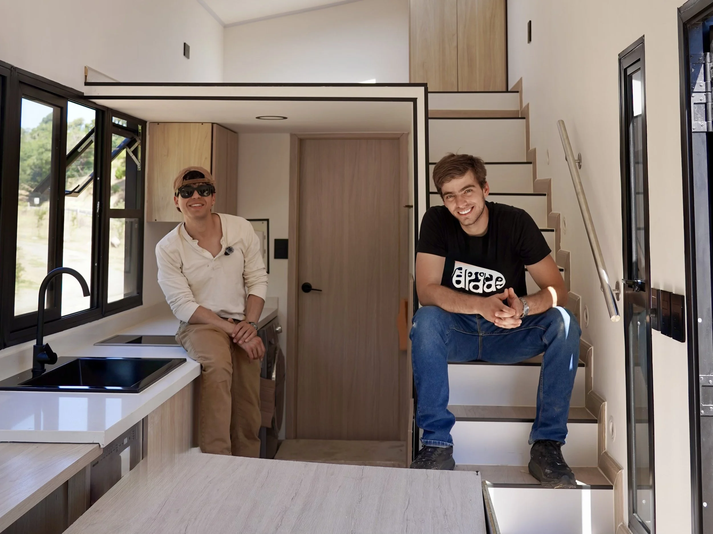 Two young men smiling inside a modern tiny home or RV with stairs, kitchen sink, and windows, during daytime.