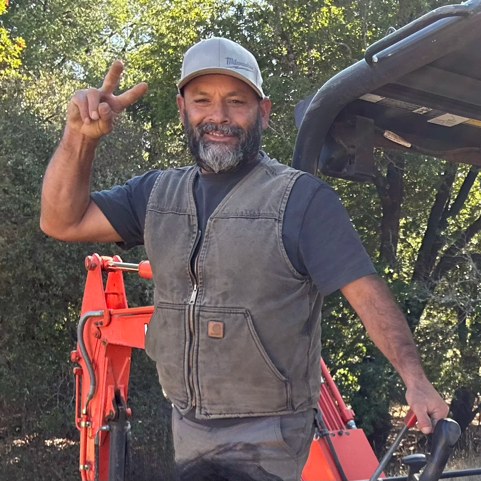 A man smiling and making a peace sign with his right hand, standing outdoors near a small orange construction vehicle, wearing a beige cap, a dark shirt, and a brown work vest, with trees in the background.