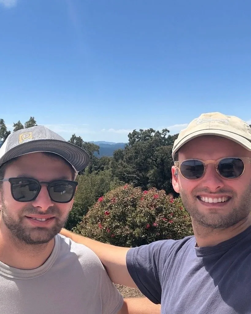 Two men are taking a selfie outdoors with a scenic background of trees and mountains. They are both wearing sunglasses and caps, and one man's arm is around the other's shoulder.