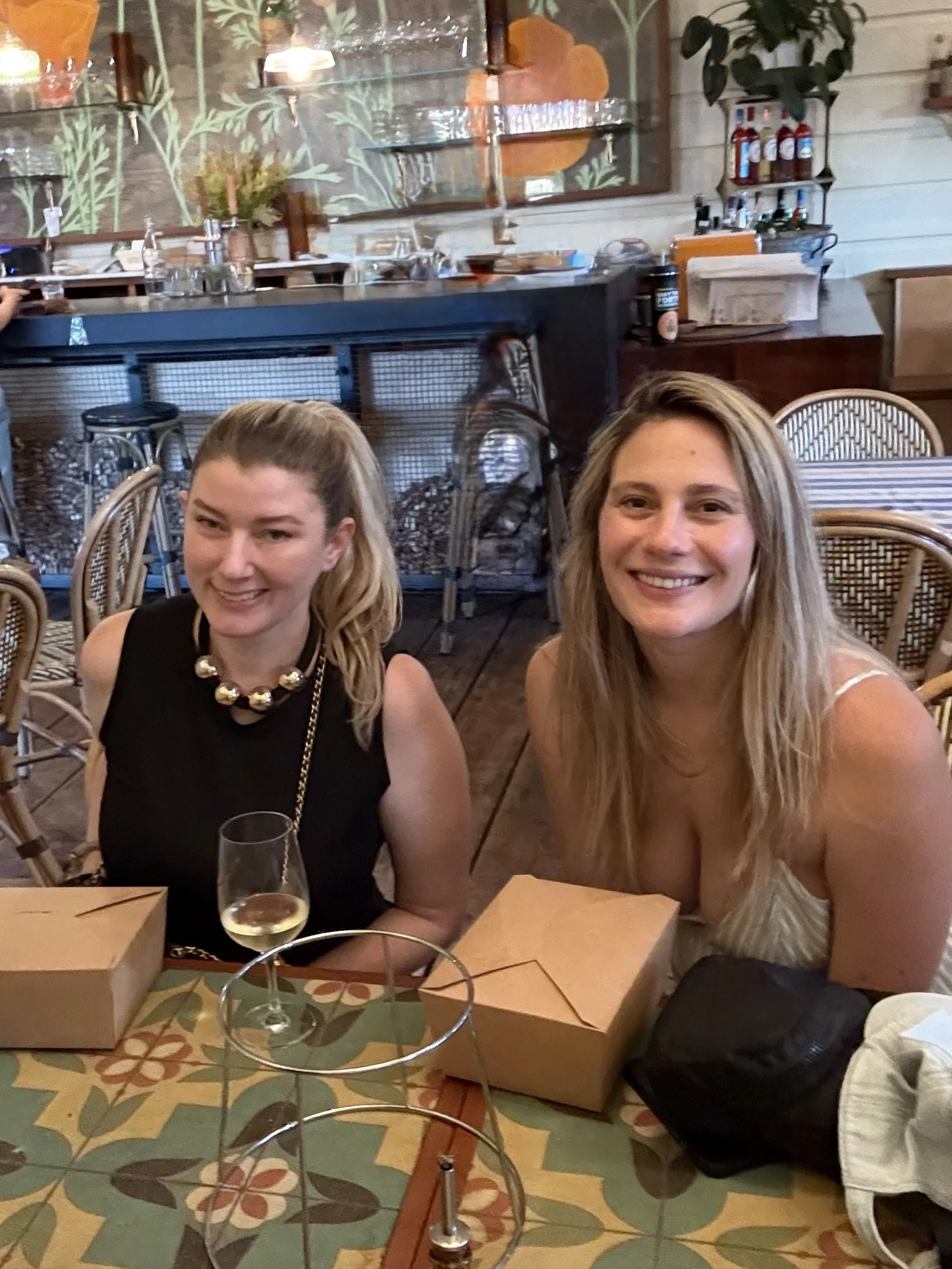 Two women sitting at a dining table in a restaurant, smiling at the camera with pizza boxes, a glass of white wine, and a black bag on the table. The restaurant has wooden walls, patterned tablecloth, and a bar in the background.
