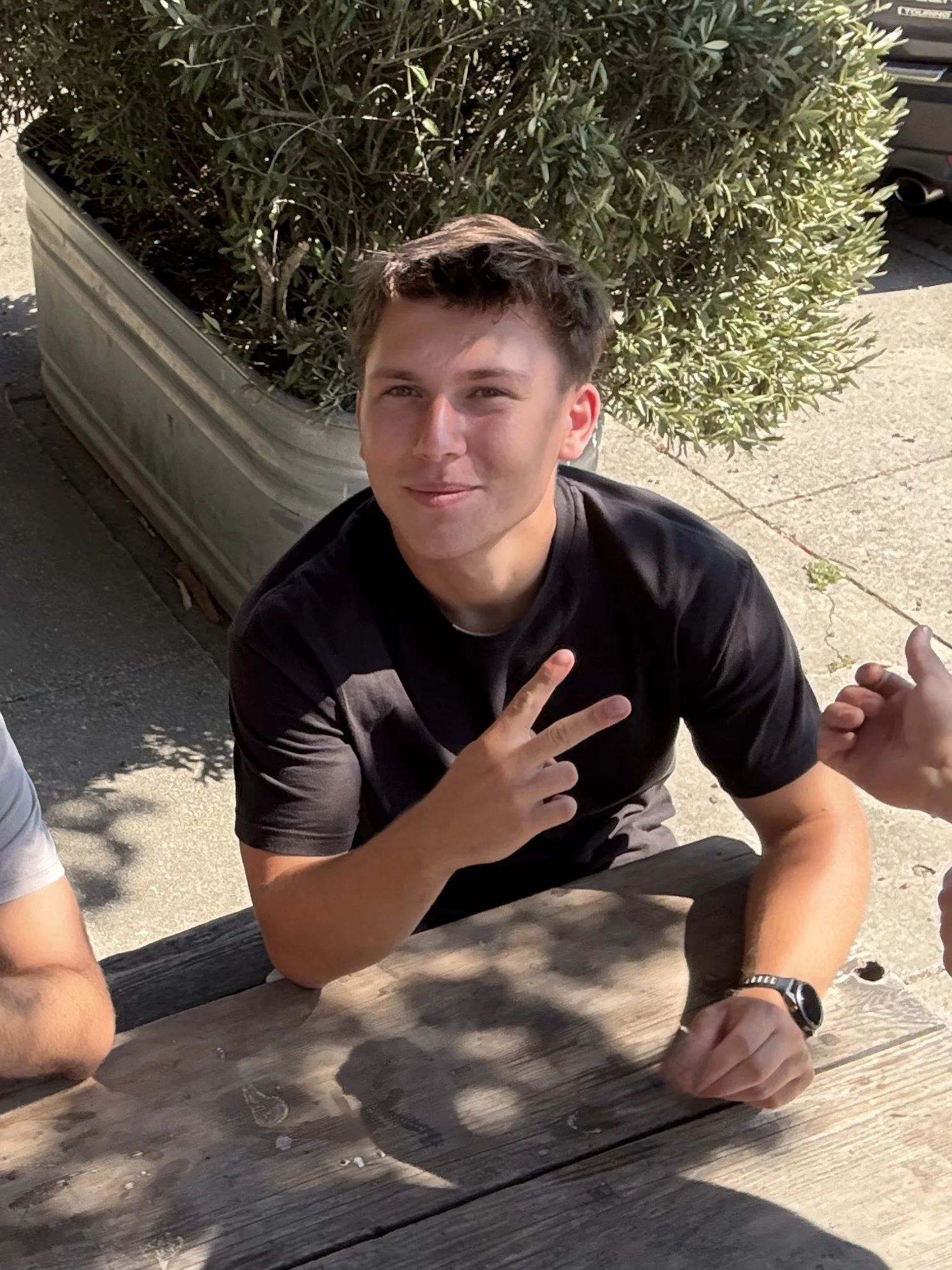 A young man sitting at a weathered wooden outdoor table, making a peace sign with his right hand. He has short, brown hair, fair skin, and is wearing a black t-shirt. Behind him is a large rectangular planter filled with green foliage, and in the background, a sidewalk and a parked gray vehicle are visible.