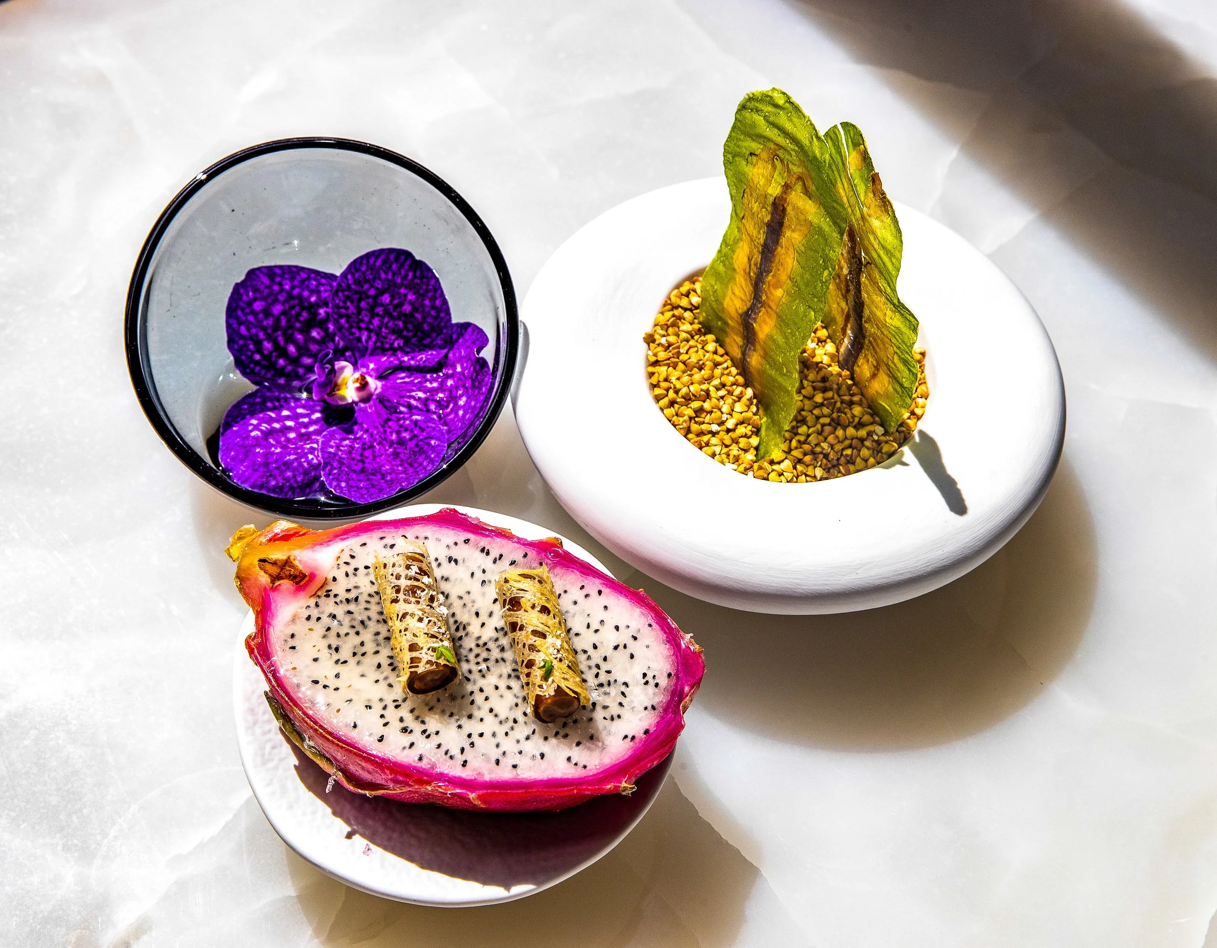 A close-up of a half-cut dragon fruit with pink outer skin and white flesh with black seeds, two rolled chocolate sticks on top, a purple orchid flower in a glass, and a potted plant with yellow pebbles and green leaves.