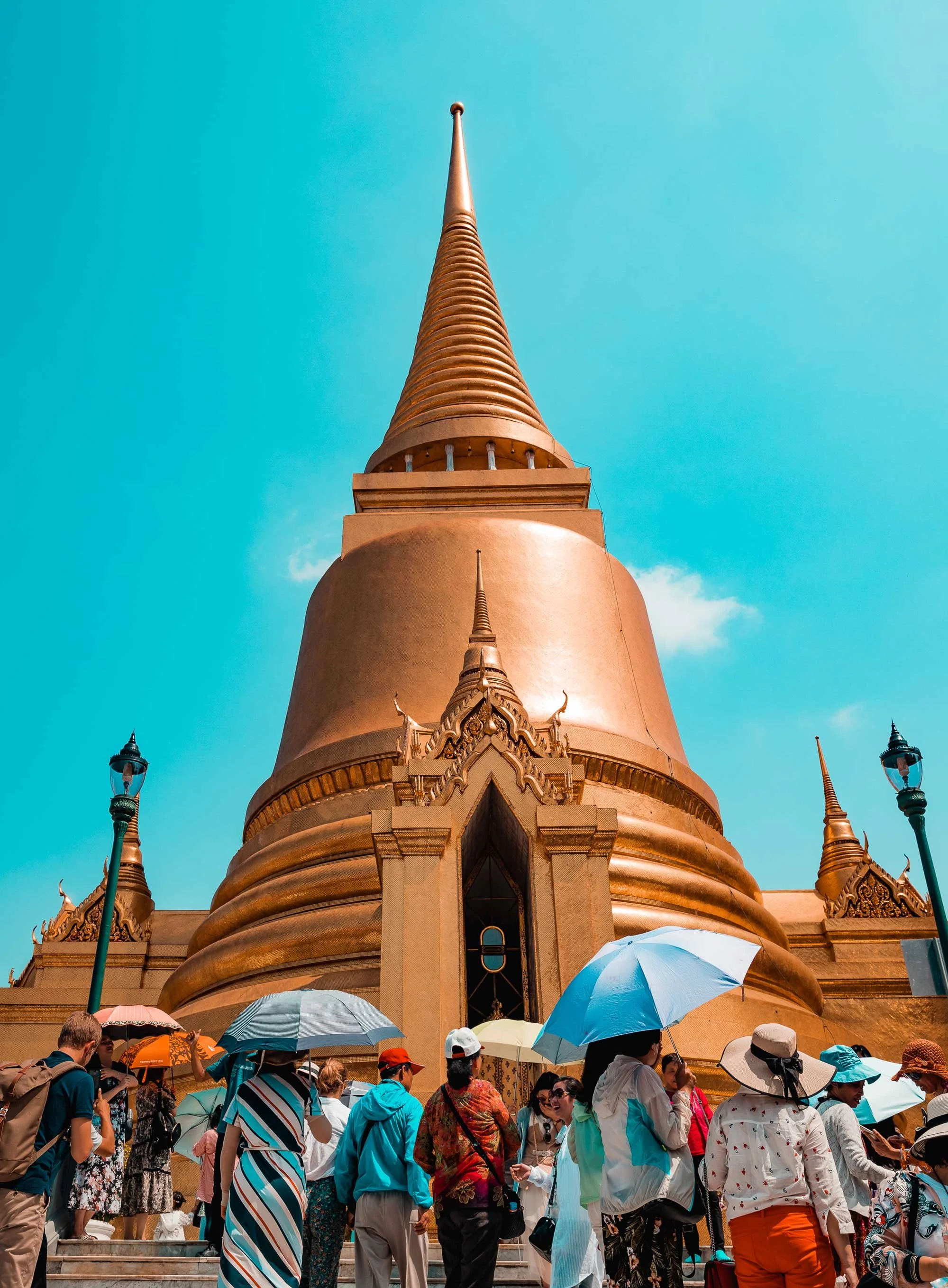 A large golden stupa or pagoda with a crowd of tourists gathered in front, many holding umbrellas, under a bright blue sky.