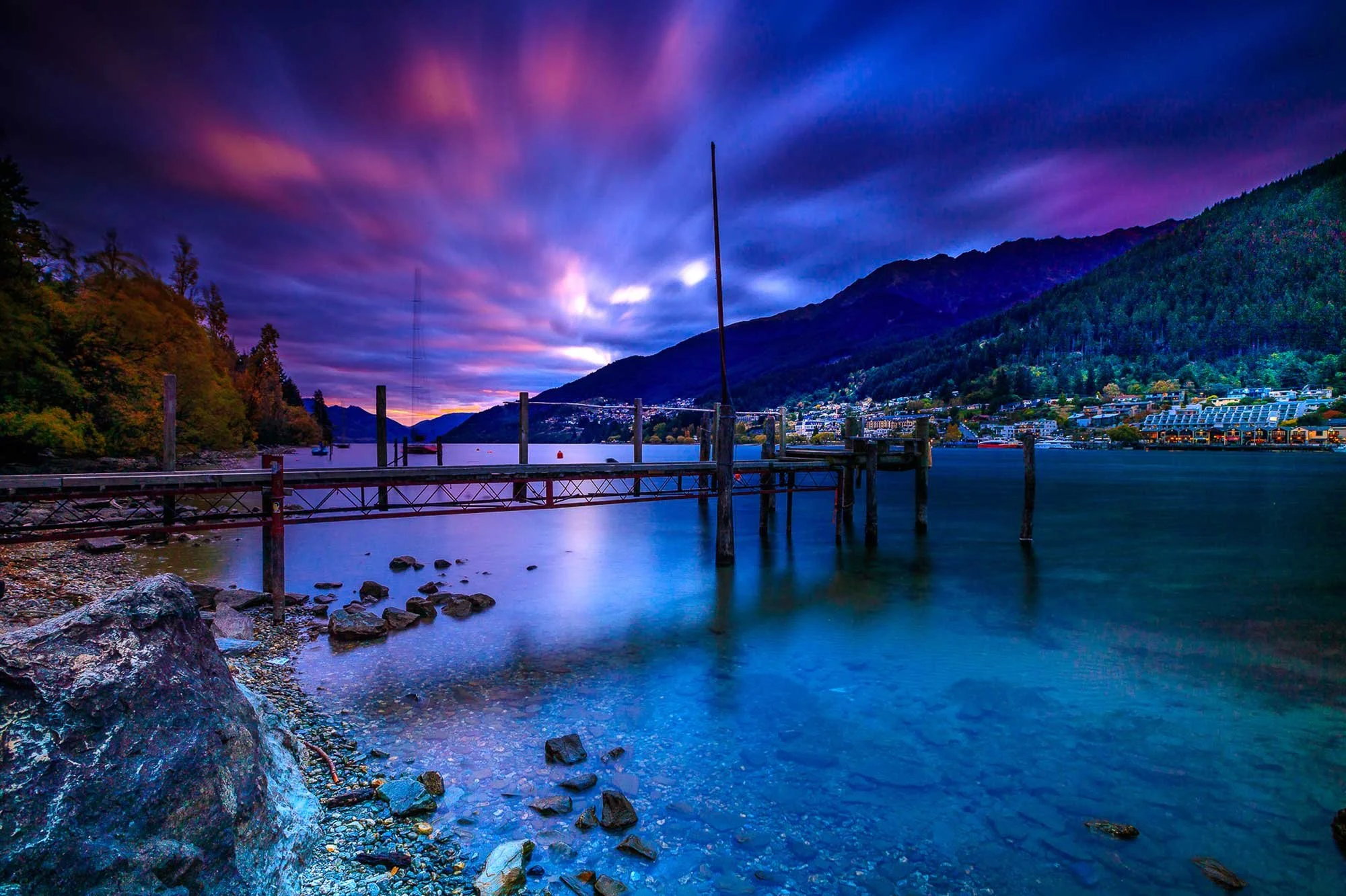 A scenic view of a river at dusk with a dock extending into the water, surrounded by mountains and trees, with colorful clouds in the sky.