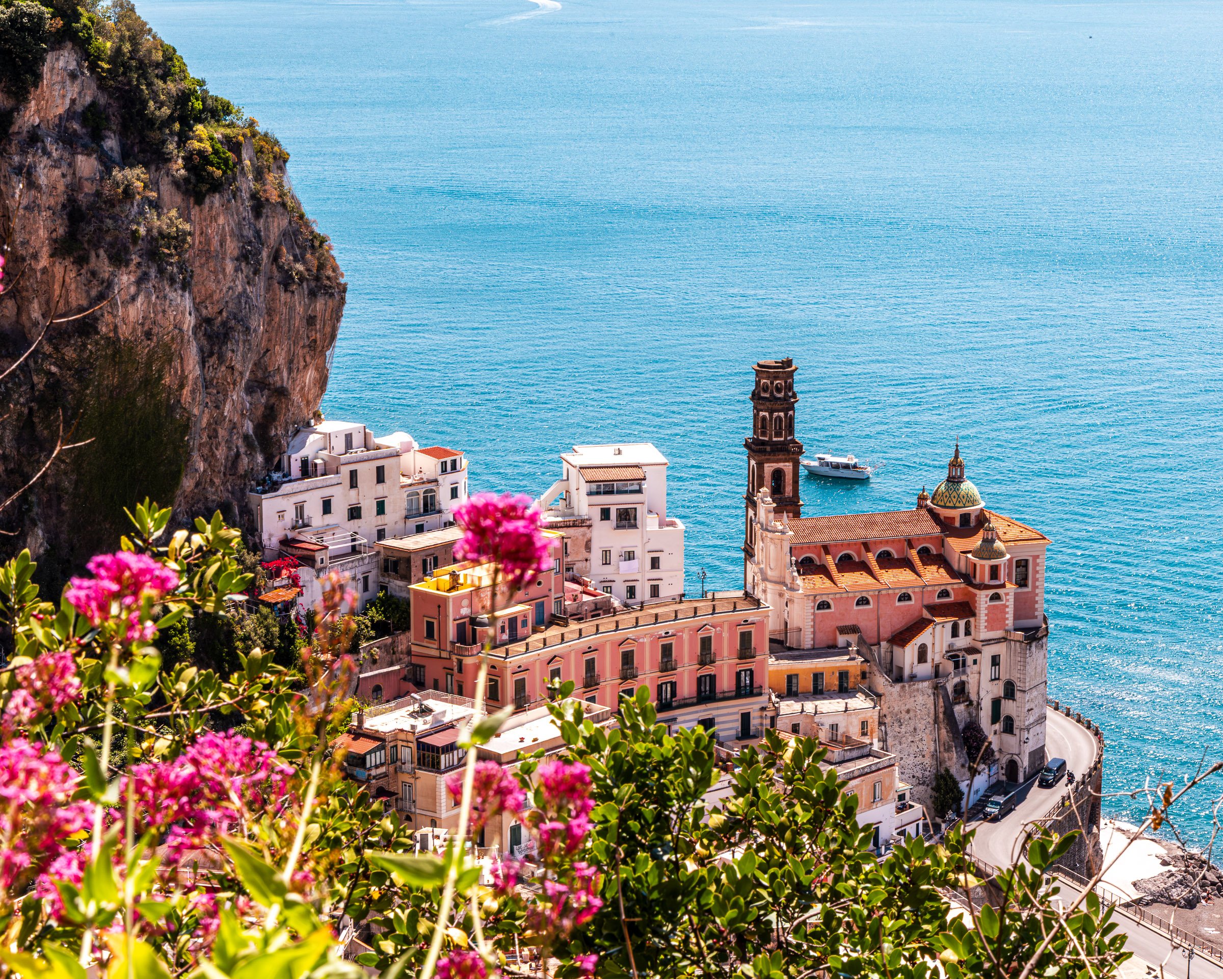 A view of a coastal town with pastel-colored buildings, a church with a bell tower, and a hillside covered in greenery overlooking the blue sea.
