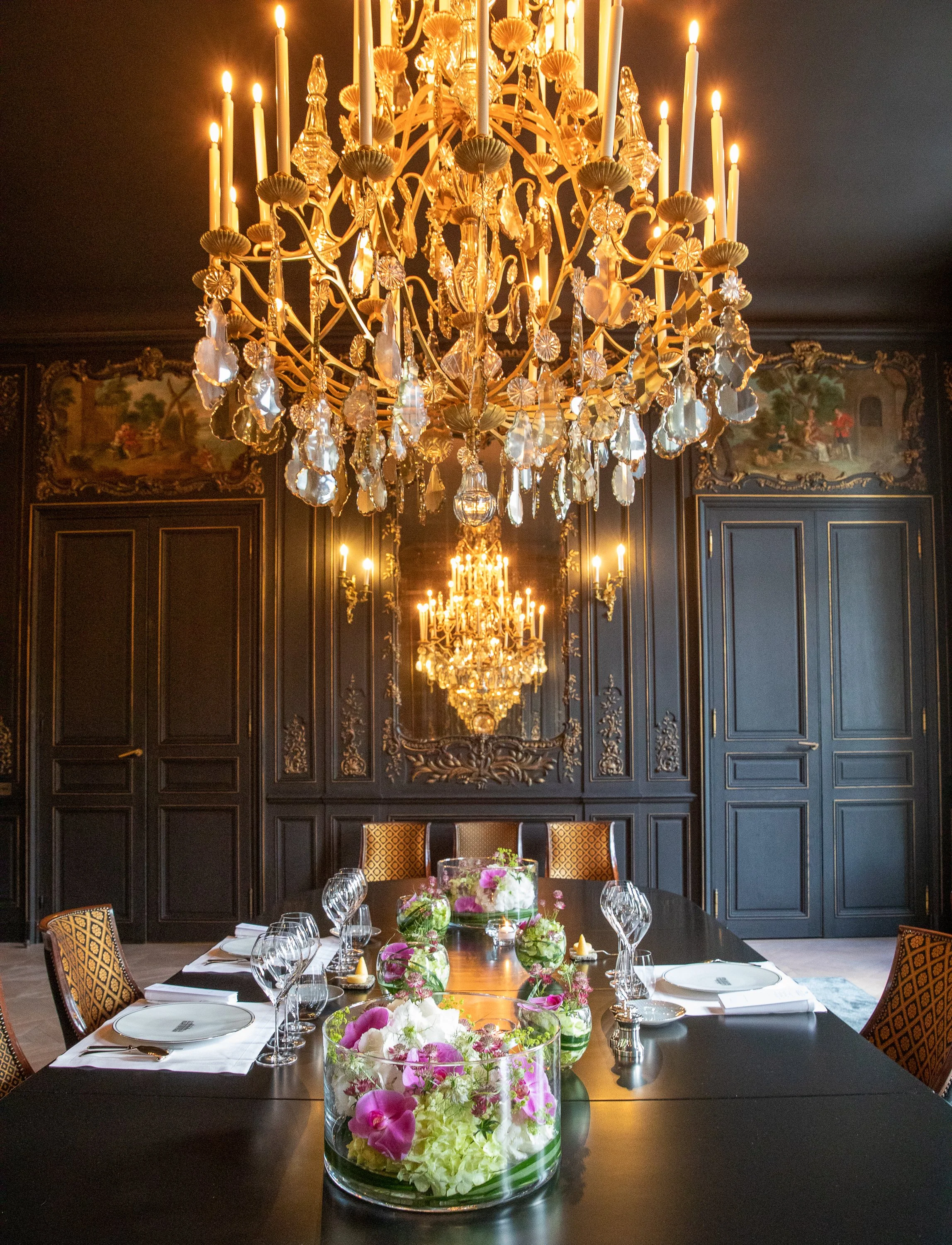 An elegant dining room with a long table set for a formal meal. The table features a centerpiece of pink, white, and green flowers in glass vases. The room has dark wood-paneled walls with ornate moldings and murals. A large, ornate crystal chandelie