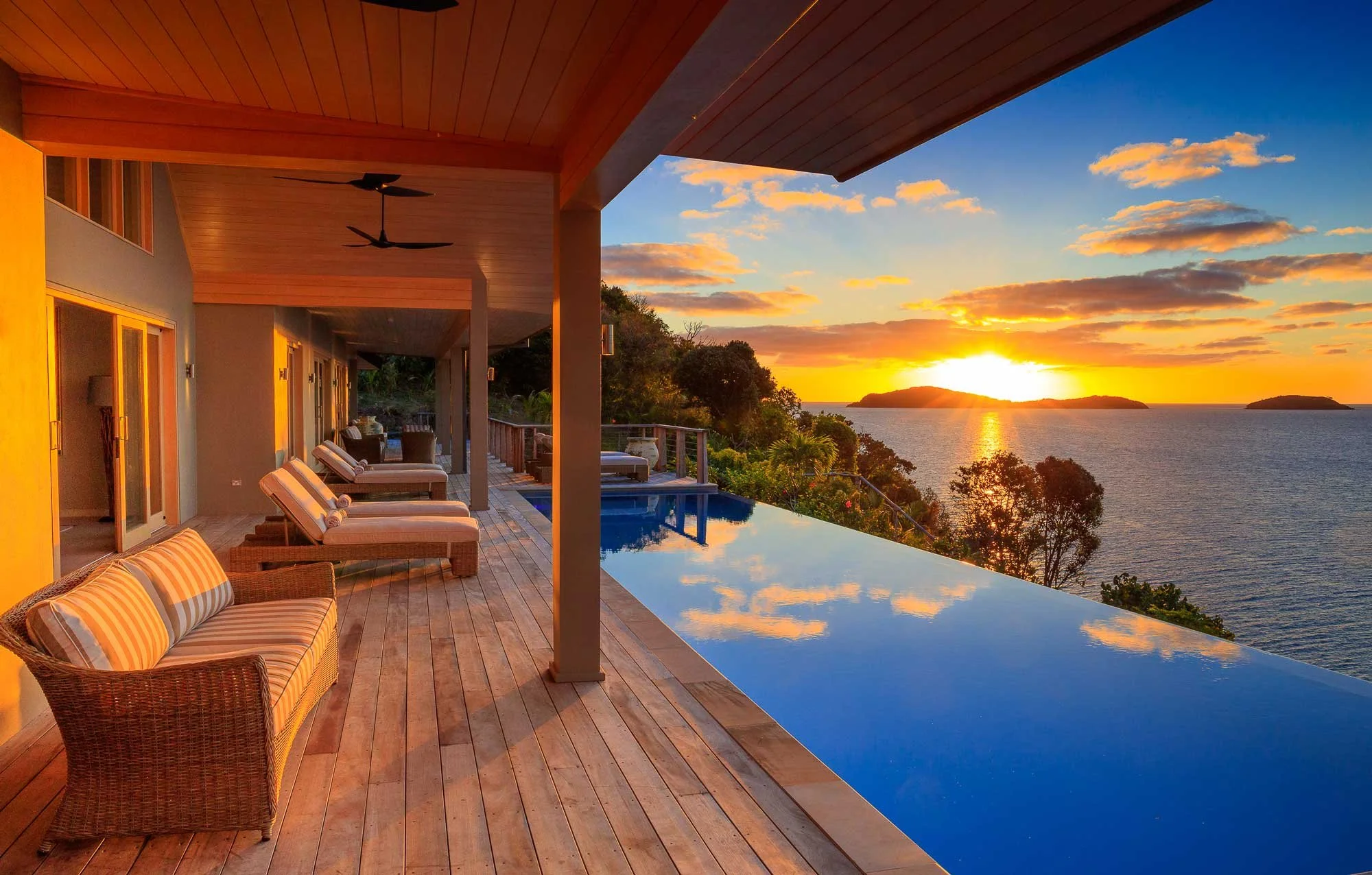 A balcony with wicker and striped cushioned chairs overlooking a sunset over the ocean. The balcony has a wooden ceiling with ceiling fans, glass doors, and a reflecting pool or hot tub. The scene is illuminated with warm orange and yellow tones from