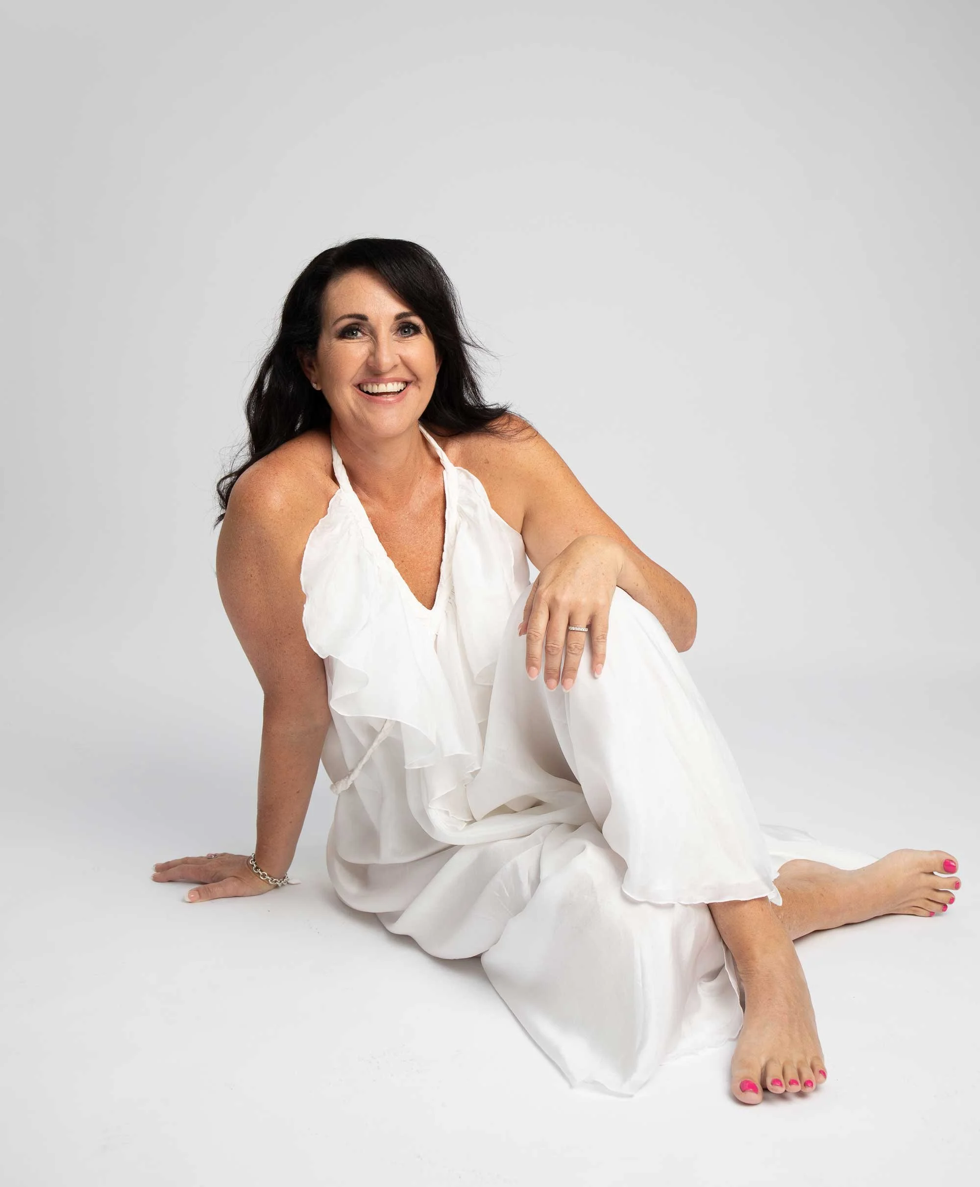 A smiling woman with dark hair wearing a white dress, sitting on the floor with her leg extended and hand resting on the ground, against a plain light gray background.