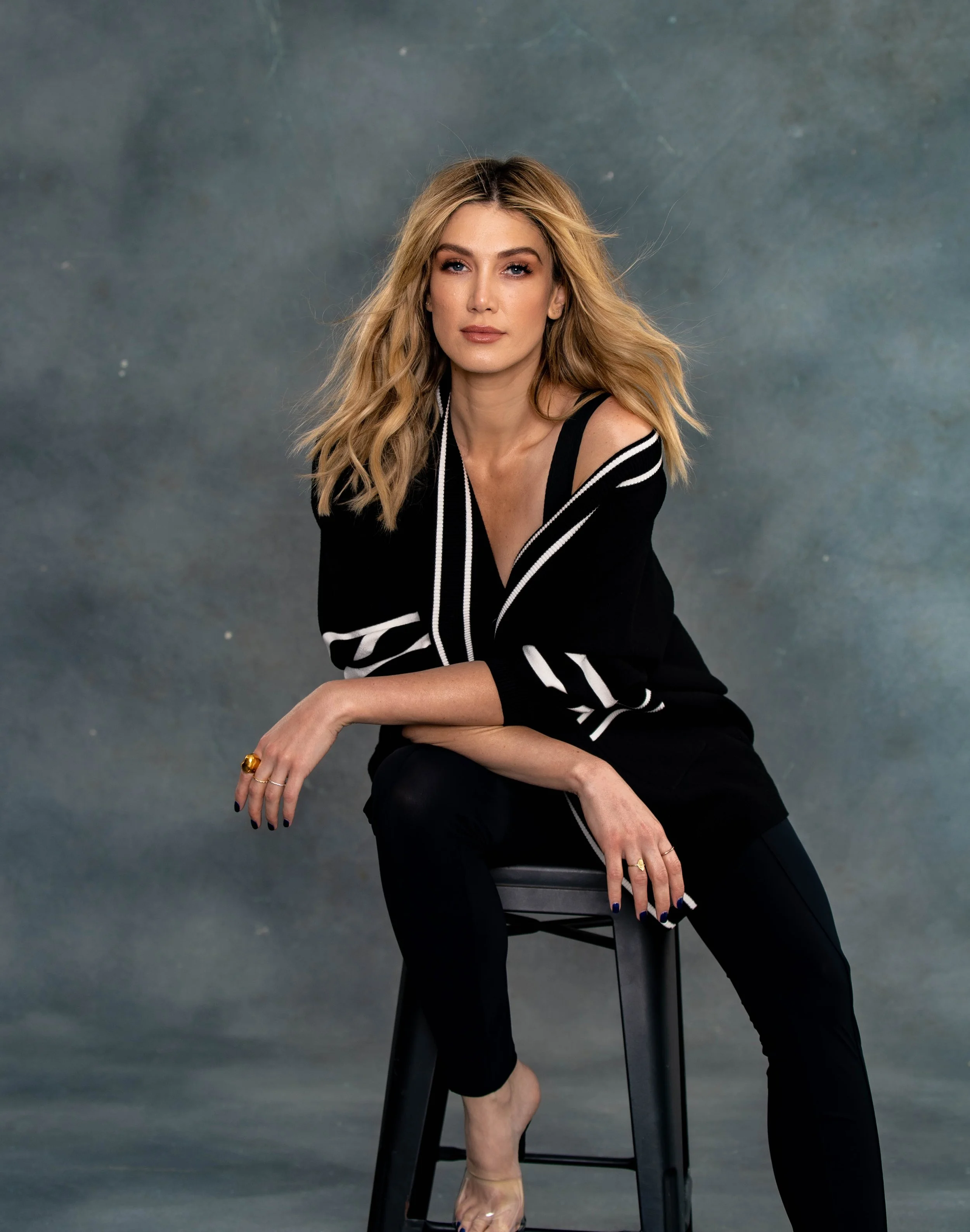 A woman with blonde, wavy hair and fair skin, dressed in a black top with white trim, is sitting on a chair with a gray background. She has a relaxed pose with one arm resting on her knee and the other on her lap, wearing rings, and looking directly at the camera.