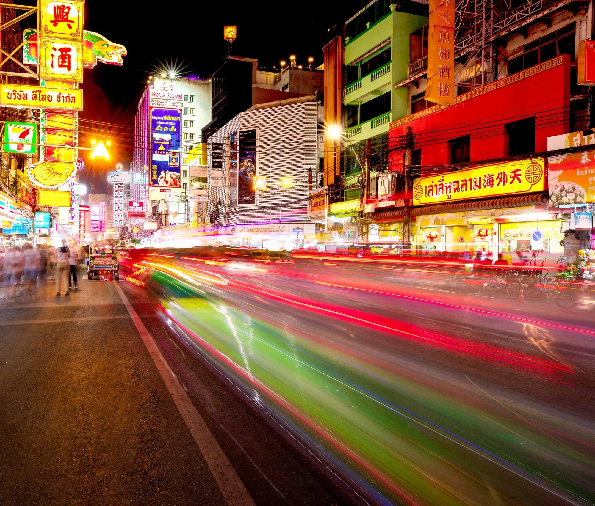 Nighttime city street scene with neon signs in Asian characters, colorful buildings, and light trails from moving vehicles.