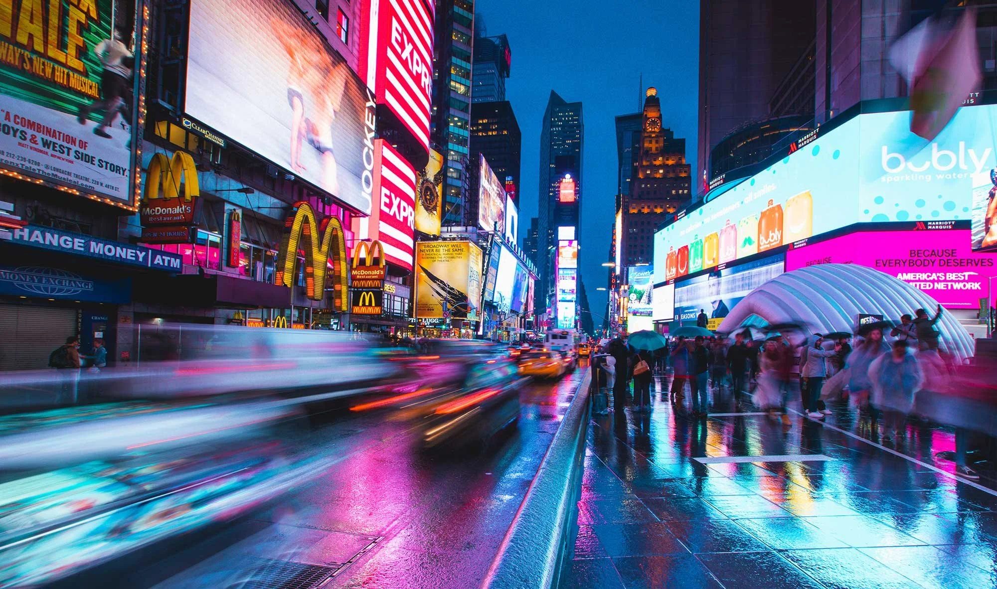 Times Square in New York City at dusk with bright digital billboards, people under umbrellas on wet pavement, and moving vehicles creating light streaks.
