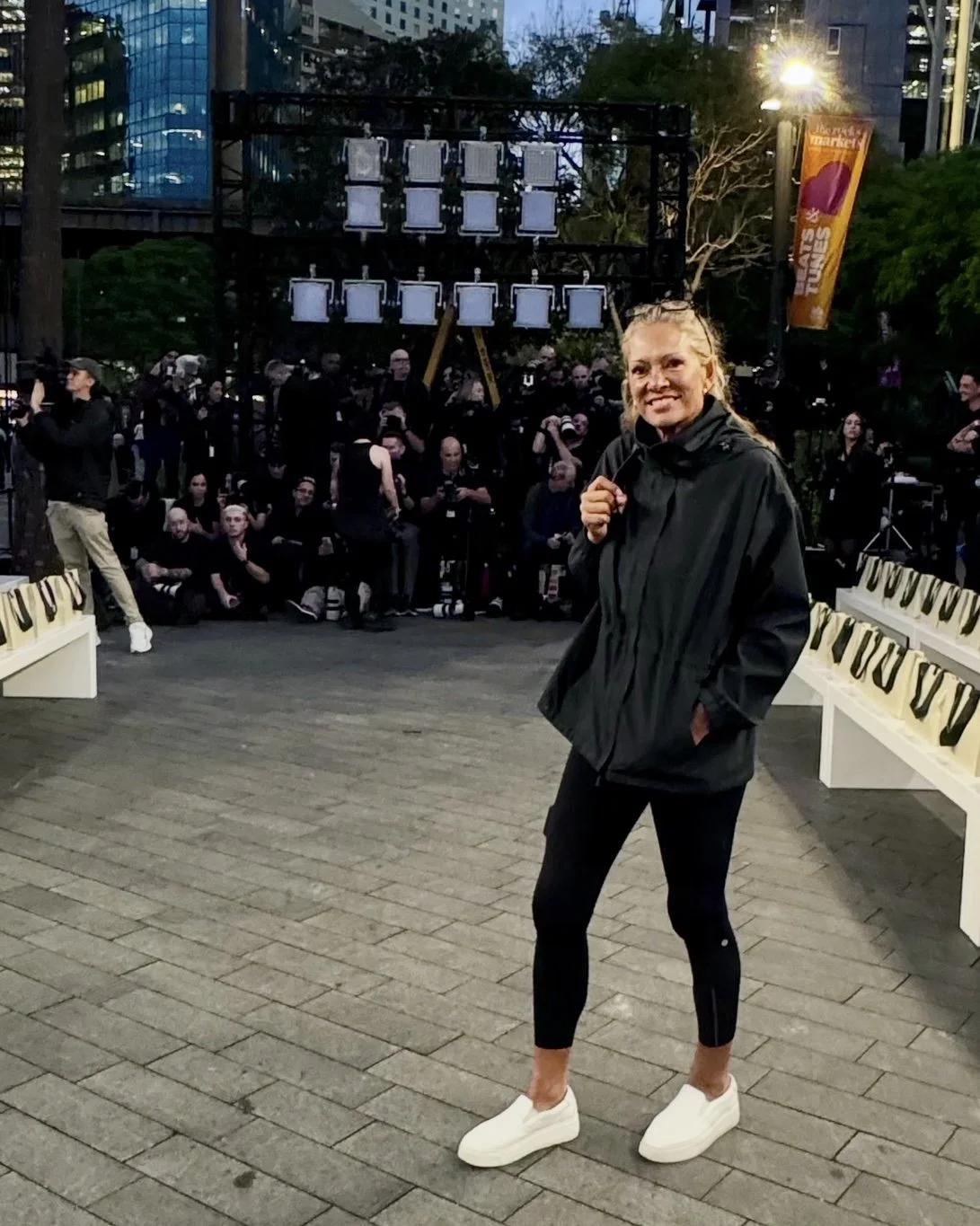 A woman in a black jacket and black leggings standing on a city street at dusk, smiling at the camera. Behind her, there are benches with letter decals, a crowd of people, and large buildings with lit windows. The scene is illuminated by streetlights and outdoor event lighting.