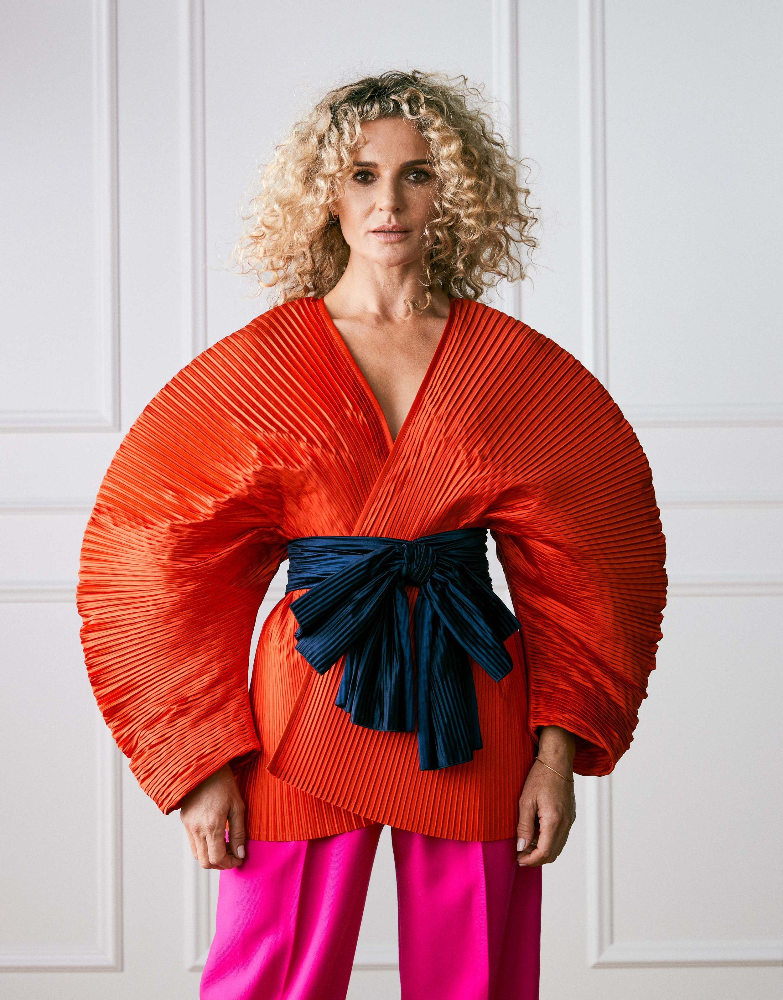 A woman with curly blonde hair wearing a red pleated top with wide sleeves, a navy blue sash tied around her waist, and bright pink pants, standing in front of a white paneled wall.