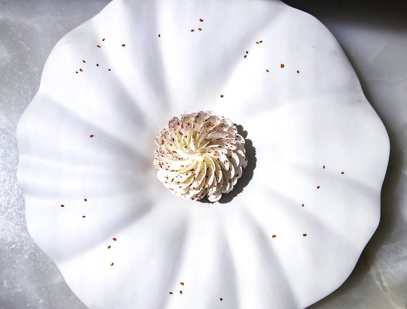 A white, ruffled flower-shaped pastry with pink sprinkles, placed on a large white plate with smaller red sprinkles around it.