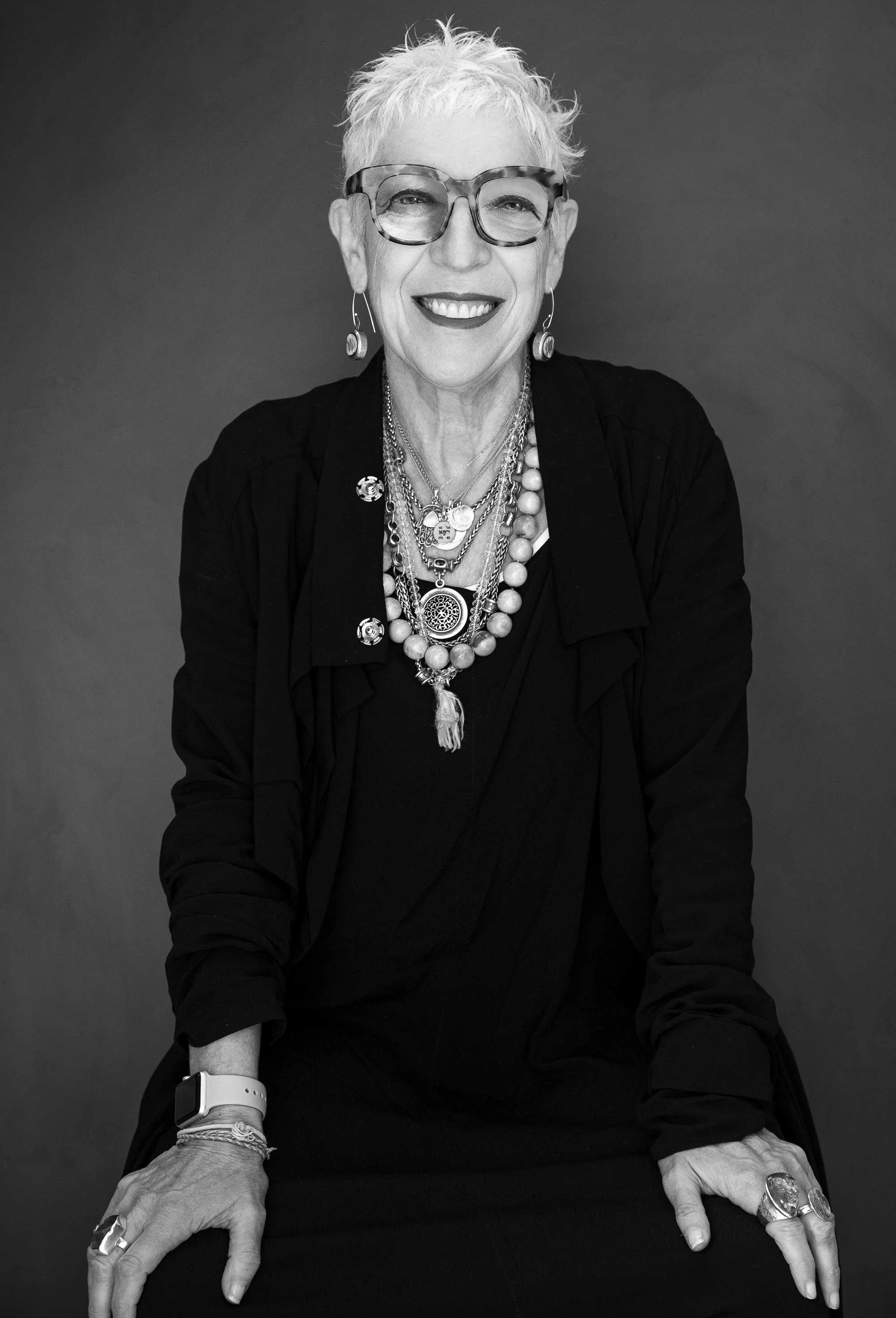 Black and white portrait of an older woman with short, tousled hair, wearing large glasses, multiple necklaces, earrings, and rings. She is smiling and sitting against a plain background.