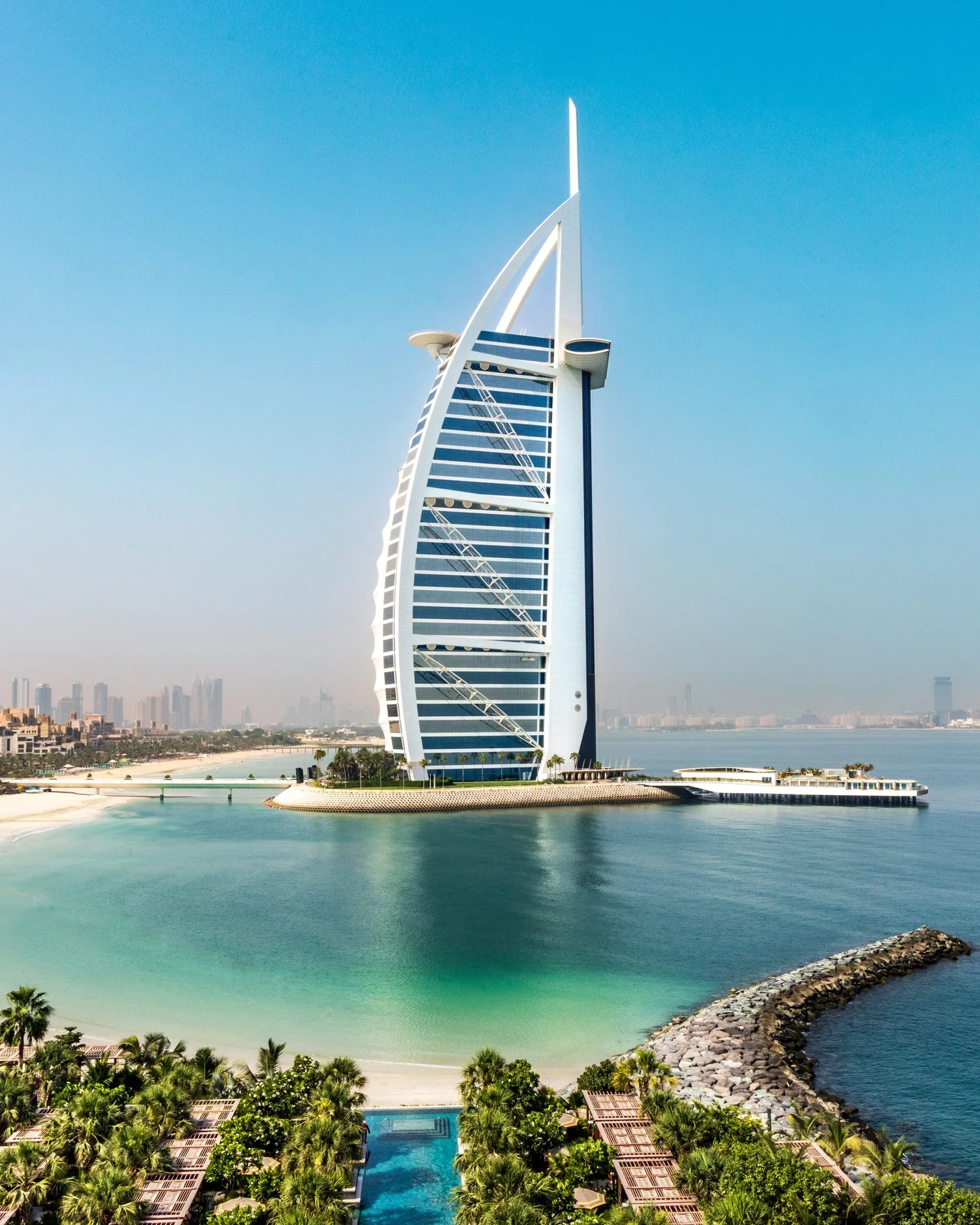 The iconic Burj Al Arab hotel, known for its sail-shaped design, stands on its own artificial island connected to the mainland by a private bridge, located in Dubai, with a clear blue sky and water surrounding it.