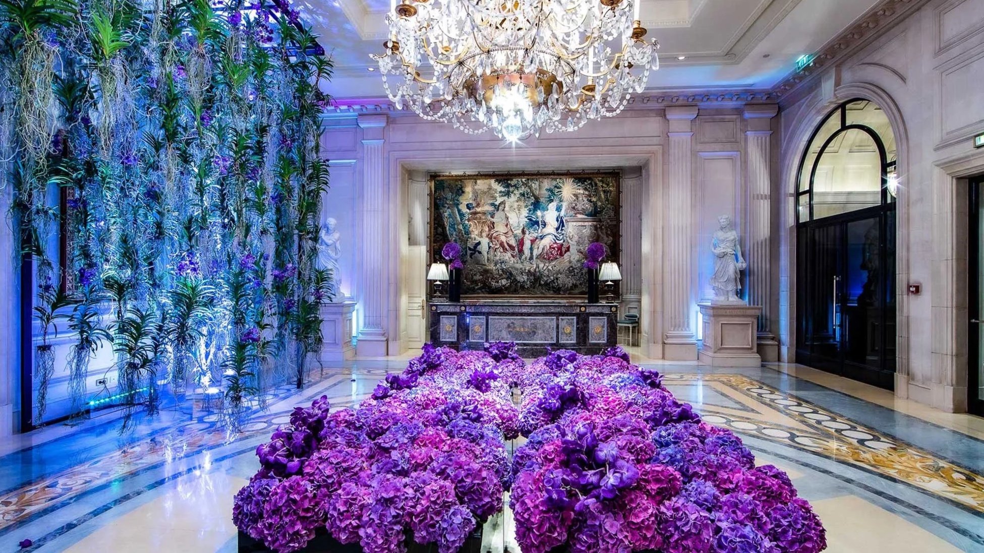 Elegant hotel lobby decorated with abundant purple flowers, a large floral arrangement on the floor, classical statues on pedestals, a decorative tapestry on the wall, ornate marble furniture, and a grand chandelier hanging from the ceiling. The space features marble floors and walls, with subtle lighting accents.