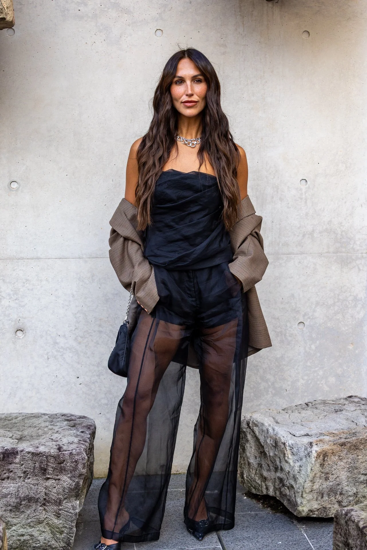 A woman with long, wavy brown hair wearing a black strapless top and sheer black wide-leg pants with brown accents, standing outdoors against a plain, light-colored concrete wall, with large rocks on either side.