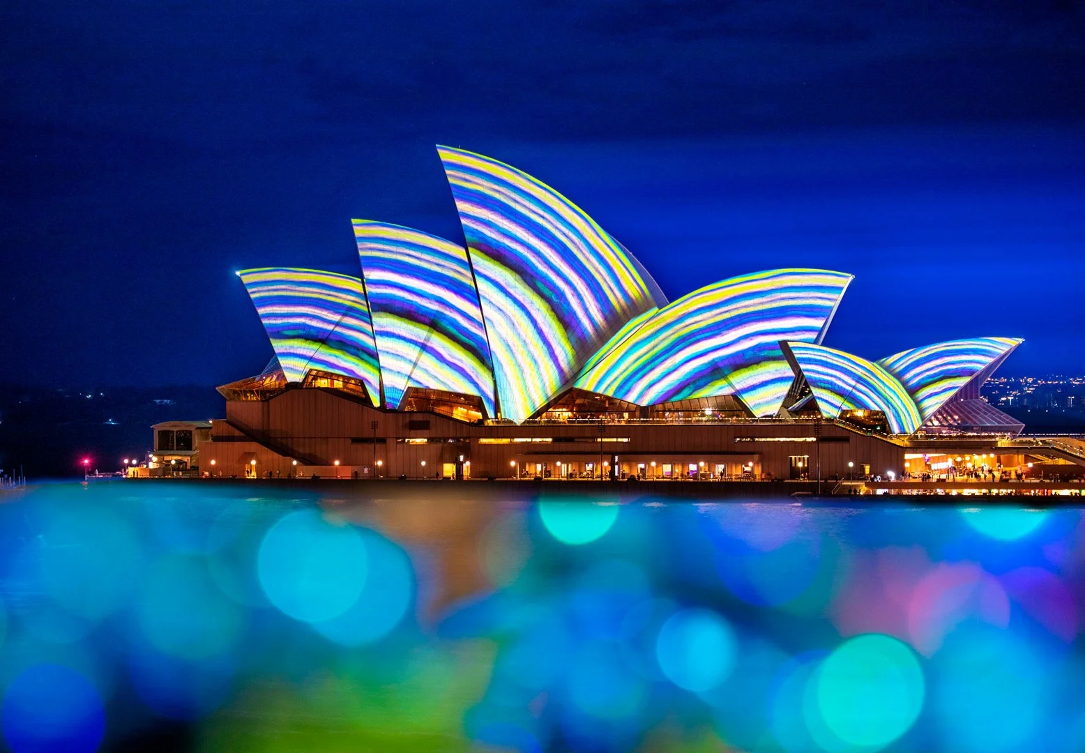 Night view of the Sydney Opera House illuminated with colorful light projections, reflecting on the water in front.