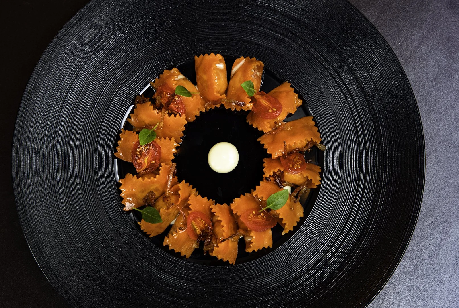 Appetizer or small dish arranged in a circle on a black textured plate, featuring folded pasta with stuffing, cherry tomato halves, small green leaves, and a small white dollop in the center.