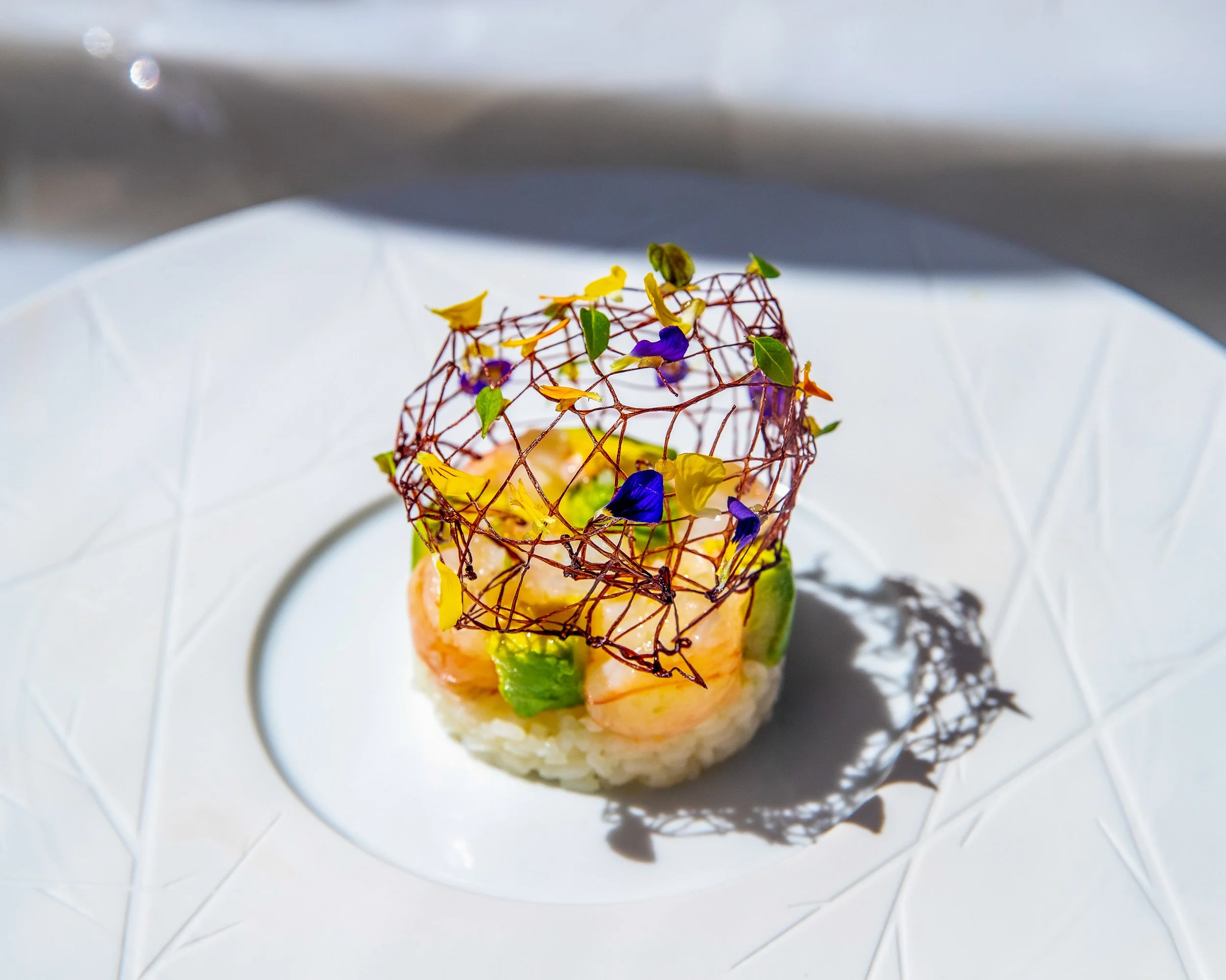 Elegant plated appetizer with shrimp, avocado, and microgreens, topped with a crispy lattice, garnished with edible flowers, drizzled with balsamic glaze, on a white plate.
