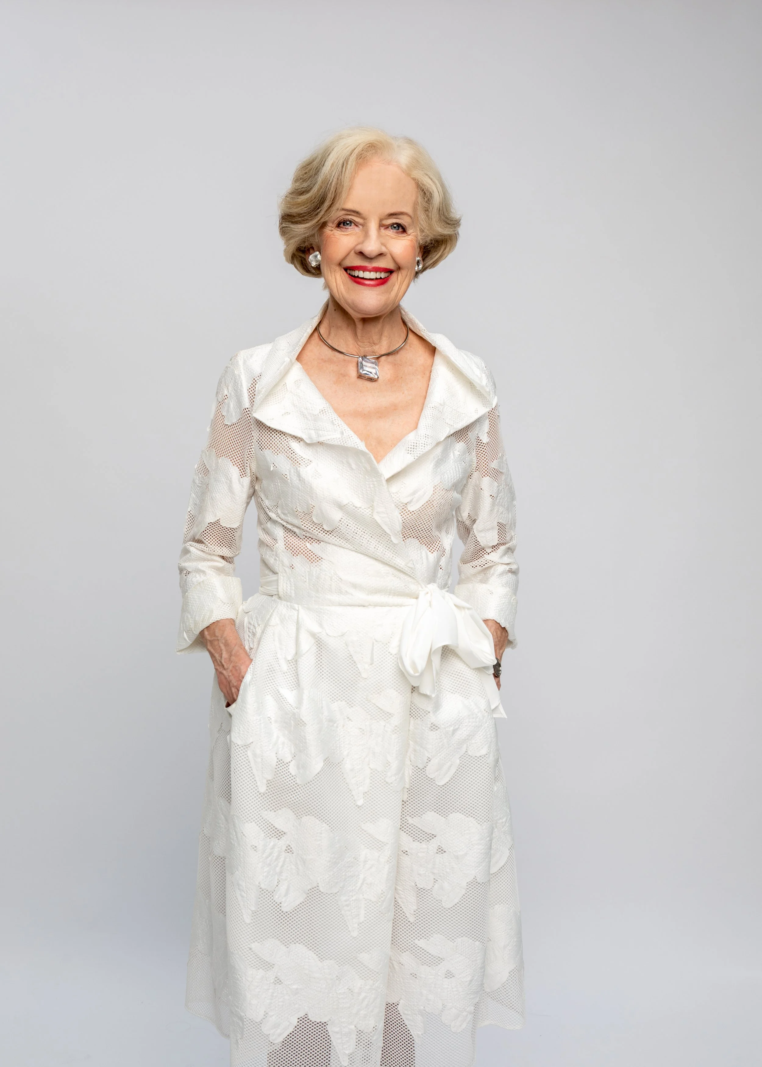 An elderly woman with short, wavy gray hair, smiling, wearing a white dress with lace details and jewelry, standing against a plain light gray background.