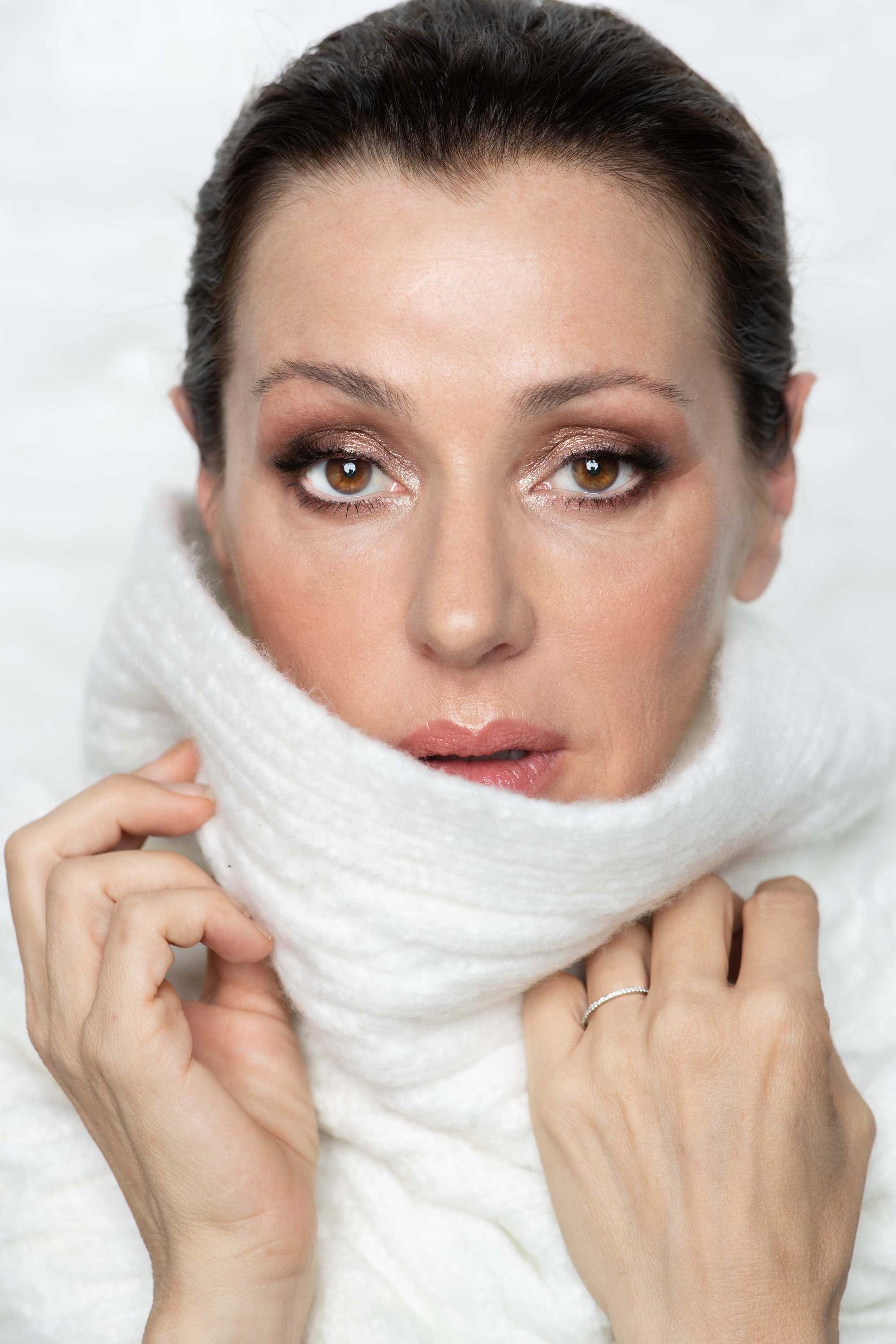 A woman with brown hair pulled back, wearing makeup, holding a white scarf close to her face, with her hands visible and wearing rings, against a white background.