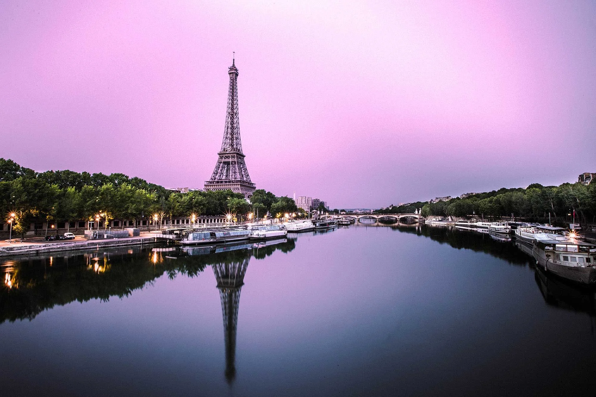 View of the Eiffel Tower over the River Seine in Paris at sunset with boats docked along the riverbank and trees lining the promenade.