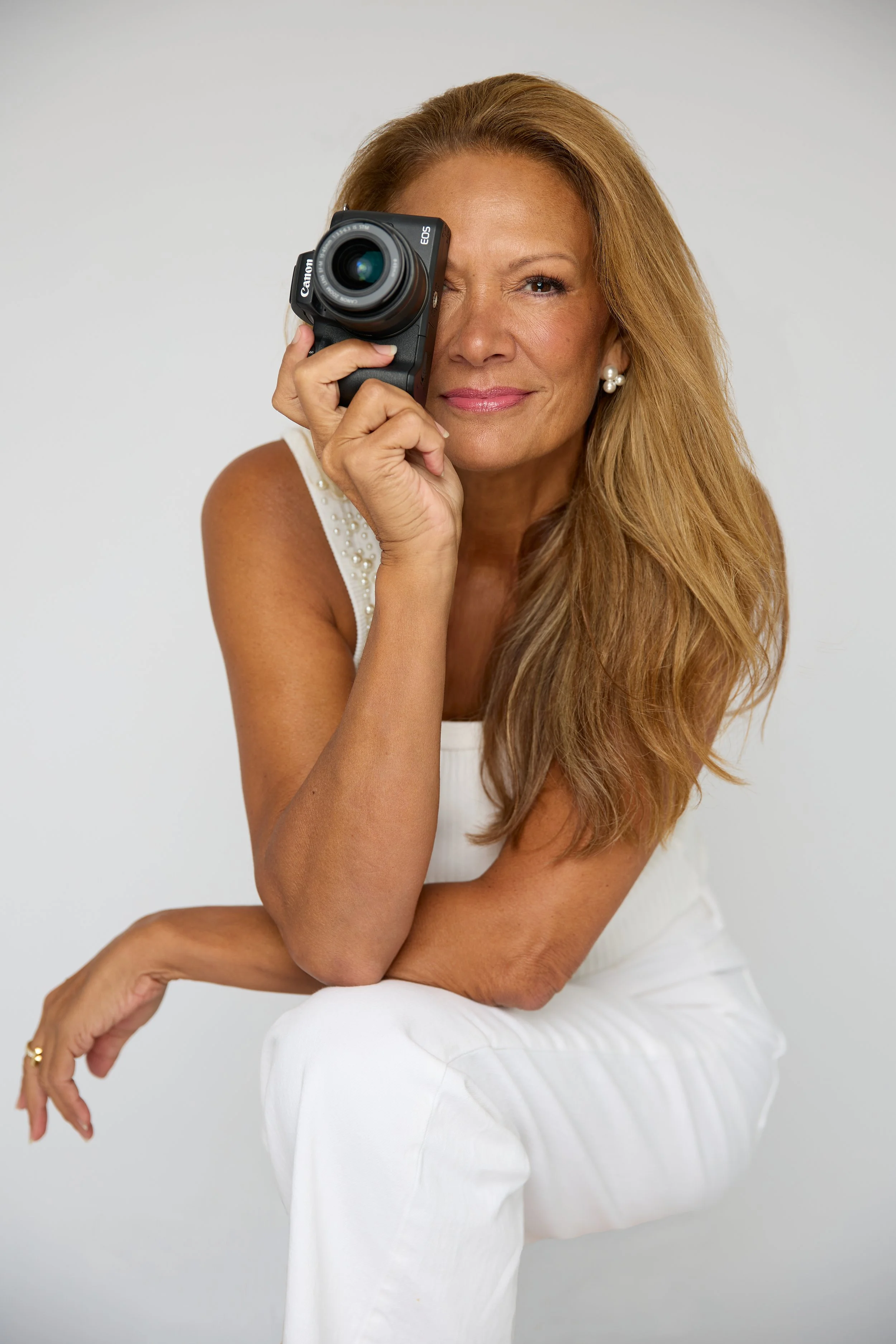 A woman with long, wavy, blonde hair in a white sleeveless top and pearl earrings, holding a camera up to her right eye, winking with a slight smile, against a plain light gray background.