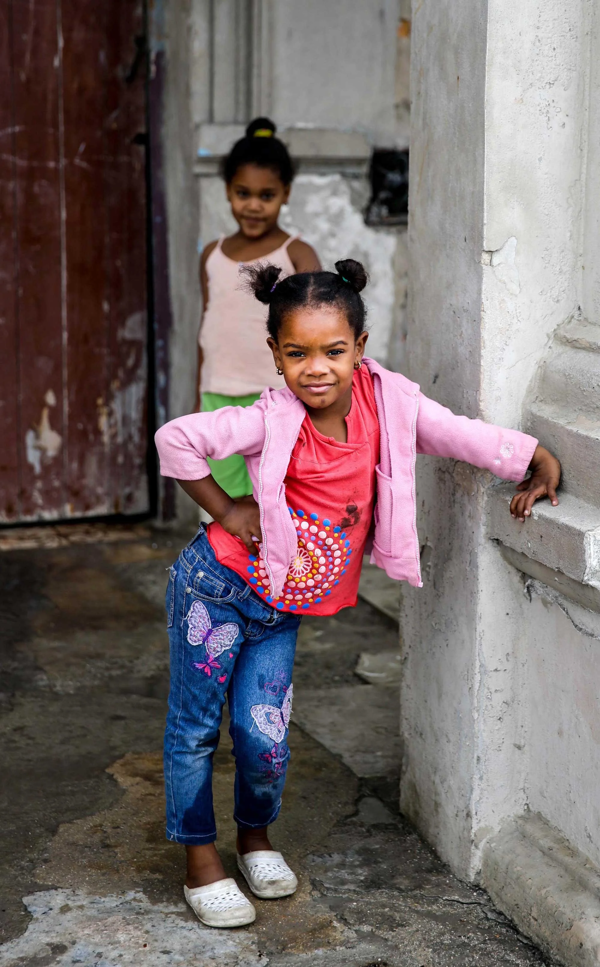 Two young girls standing outdoors near a wall made of unfinished concrete. The girl in the front wears a pink jacket, red shirt with designs, blue jeans with butterfly embroidery, and white shoes. The girl in the background wears a light pink top and